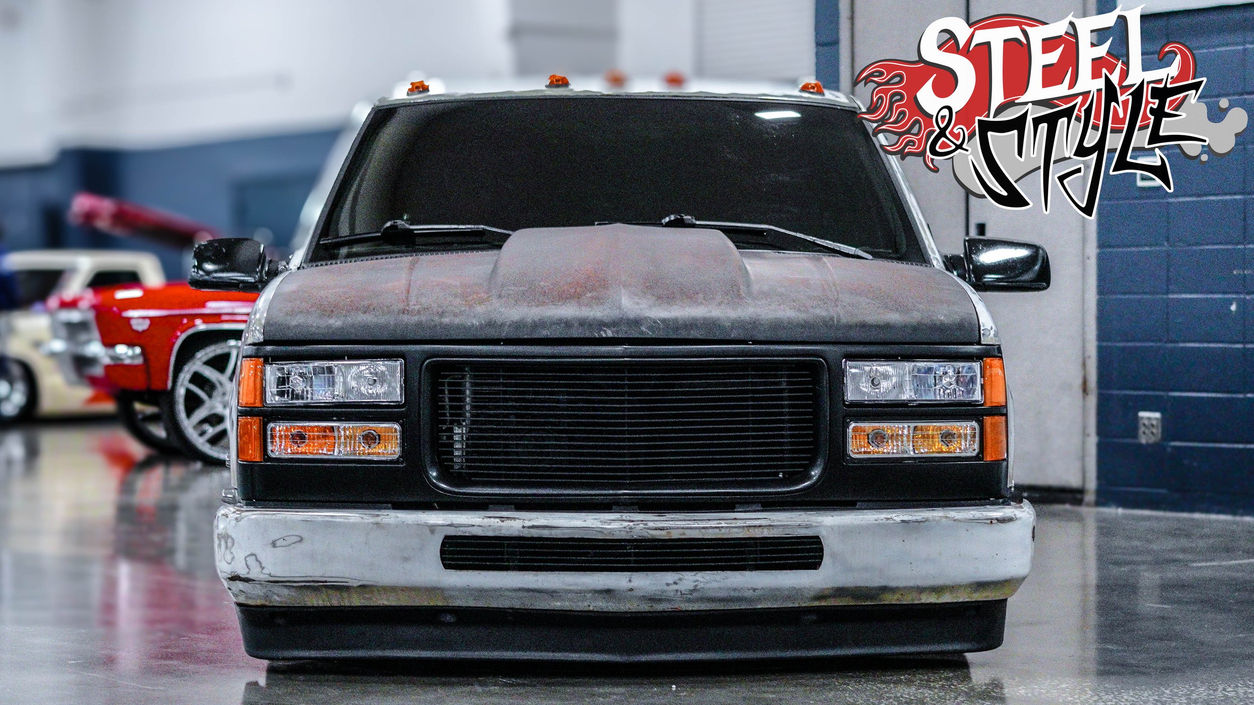 Front view of a vintage black SUV with a rusty hood, parked on a polished floor in an indoor garage with other classic cars in the background. The garage features blue and gray walls. A stylized logo reading 'STEEL & STYLE' with flames is in the uppe