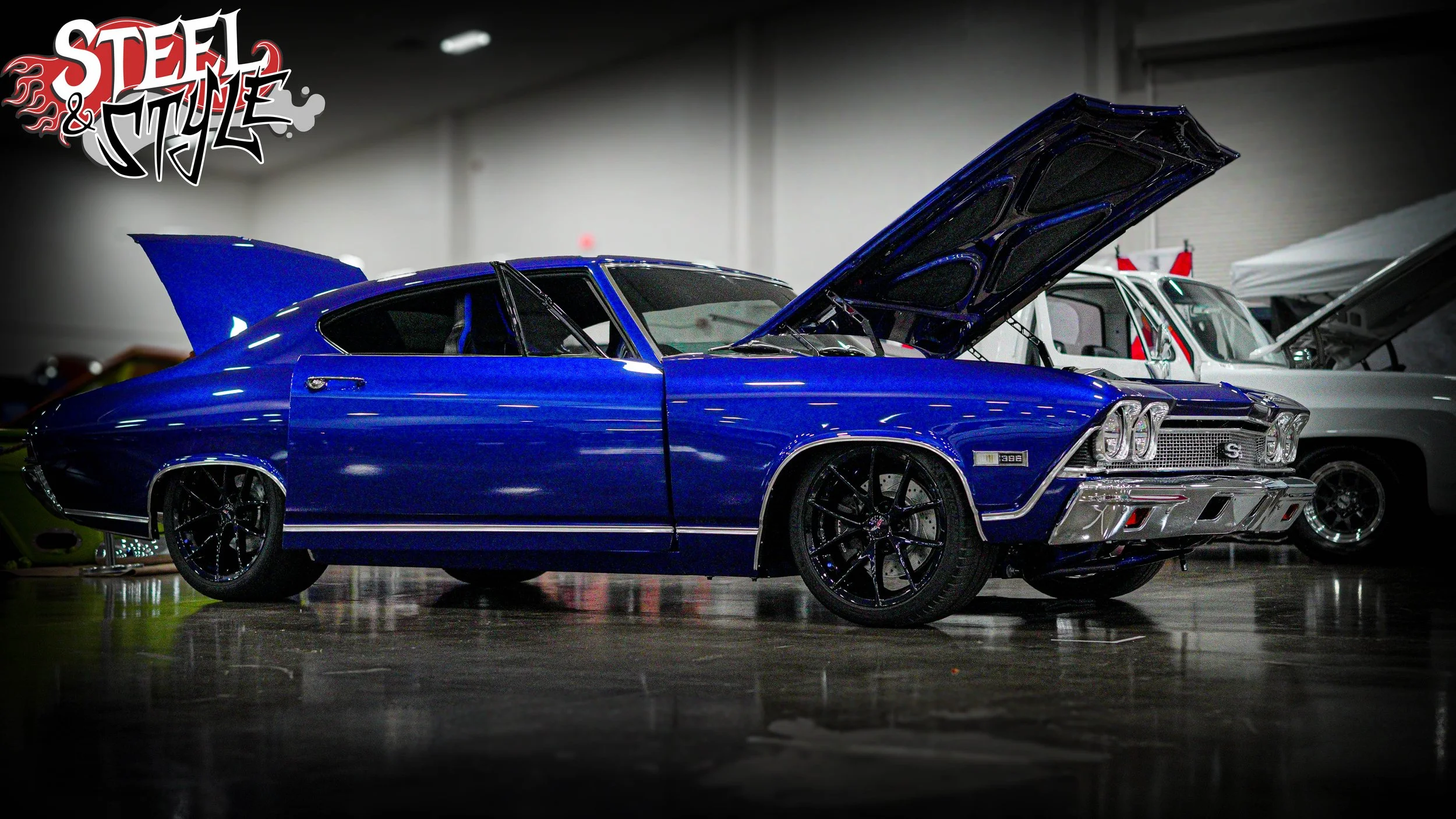 Blue classic muscle car at an indoor car show with other vehicles visible in the background, hood open, with the "Steel & Style" logo in the top left corner.