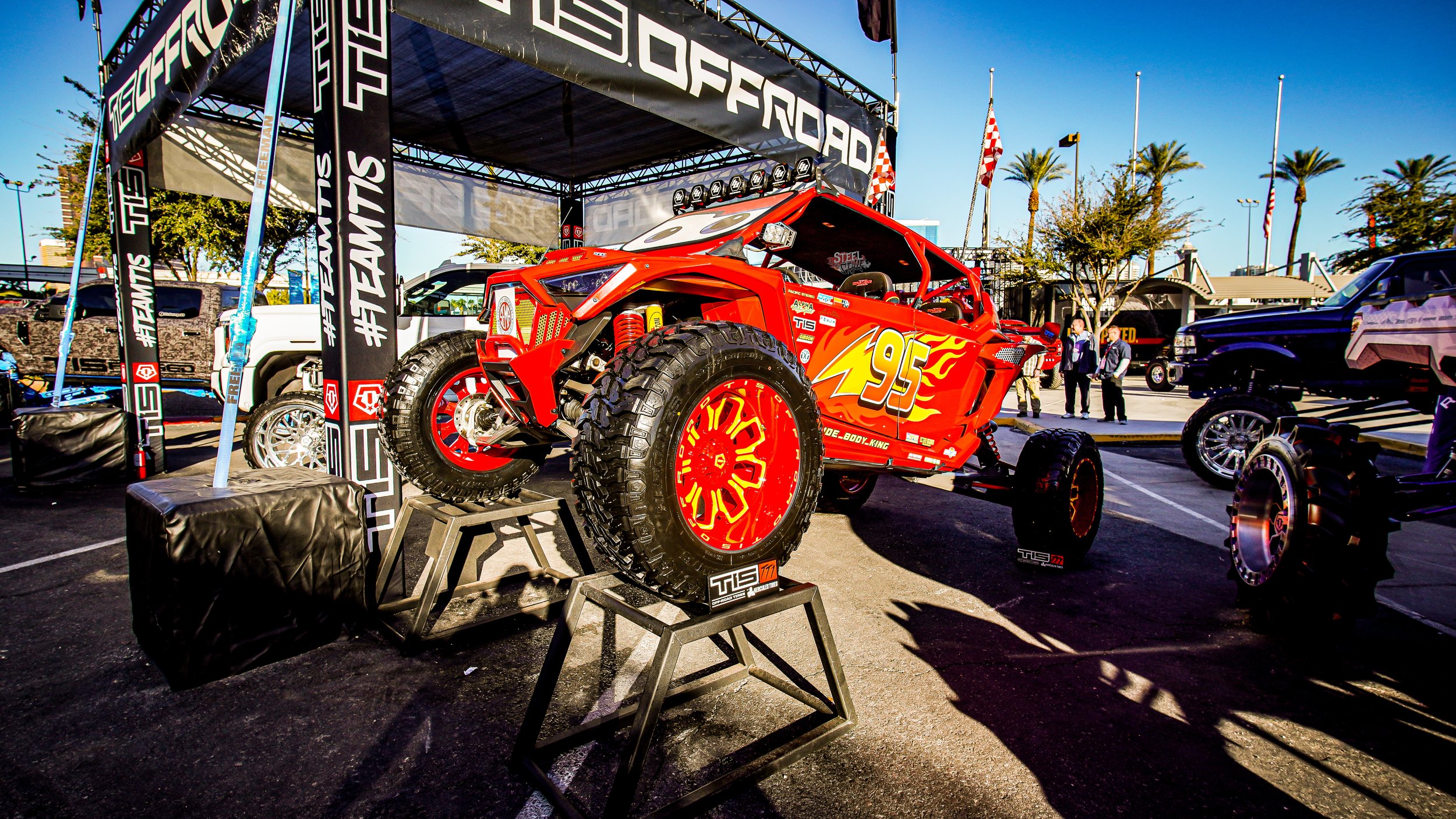 A red and yellow race car, designed like Lightning McQueen from Pixar's Cars, displayed on a stand at an outdoor event with other vehicles and people in the background.