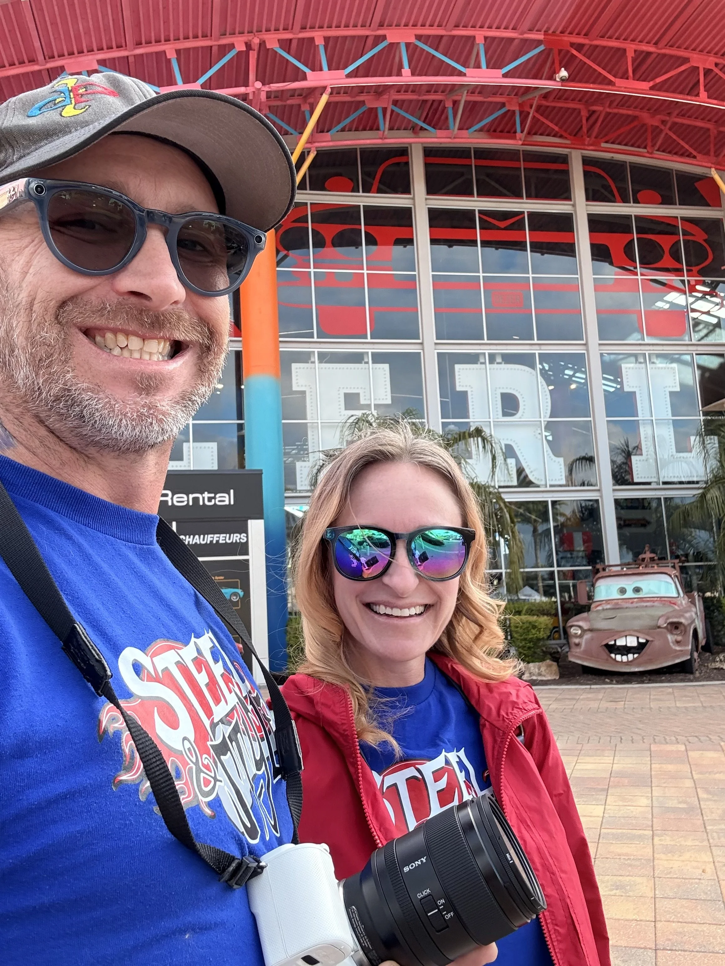 Two smiling people, a man and a woman, taking a selfie outside a building with colorful glass and a vintage car in the background. The man is wearing sunglasses, a gray cap, a blue shirt, and has a camera hanging around his neck. The woman is wearing sunglasses, a red jacket, and a blue shirt with a logo.