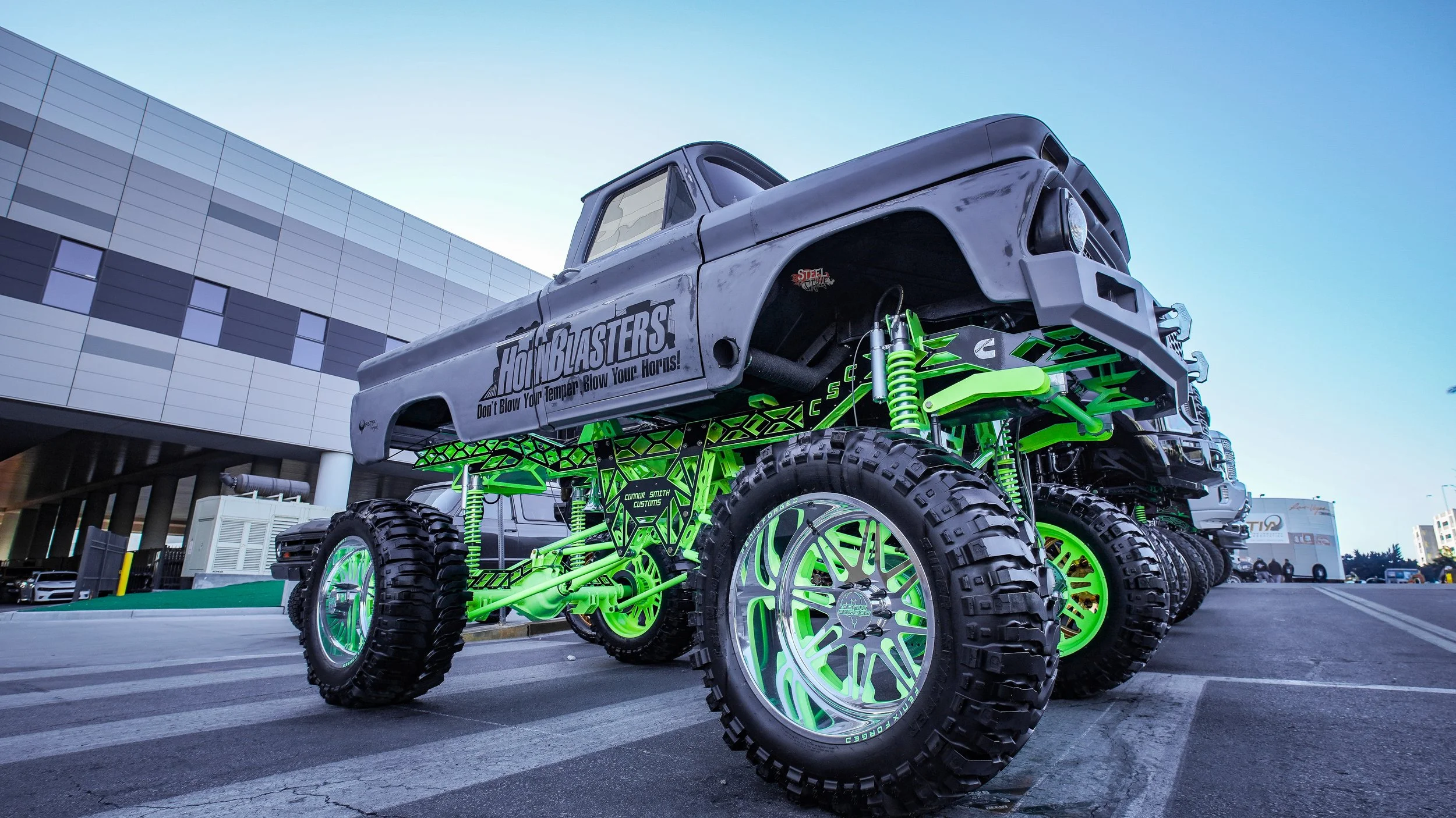 A heavily modified monster truck with large off-road tires, bright green suspension components, and a gray truck body with a decal that reads "Honk Blasters!" parked in an urban area near a modern building.
