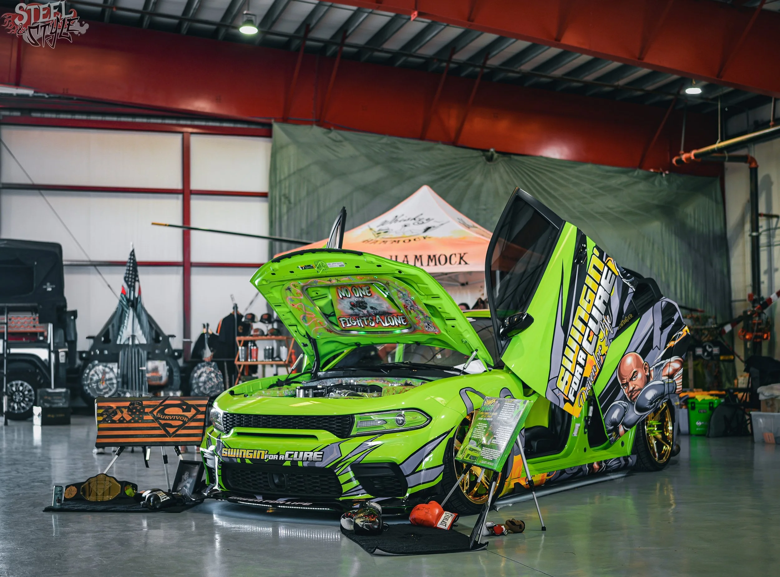 Green race car with racing decals and open hood displayed indoors, with boxing gloves, a boxing glove display stand, and memorabilia nearby, in a large warehouse or exhibition space.