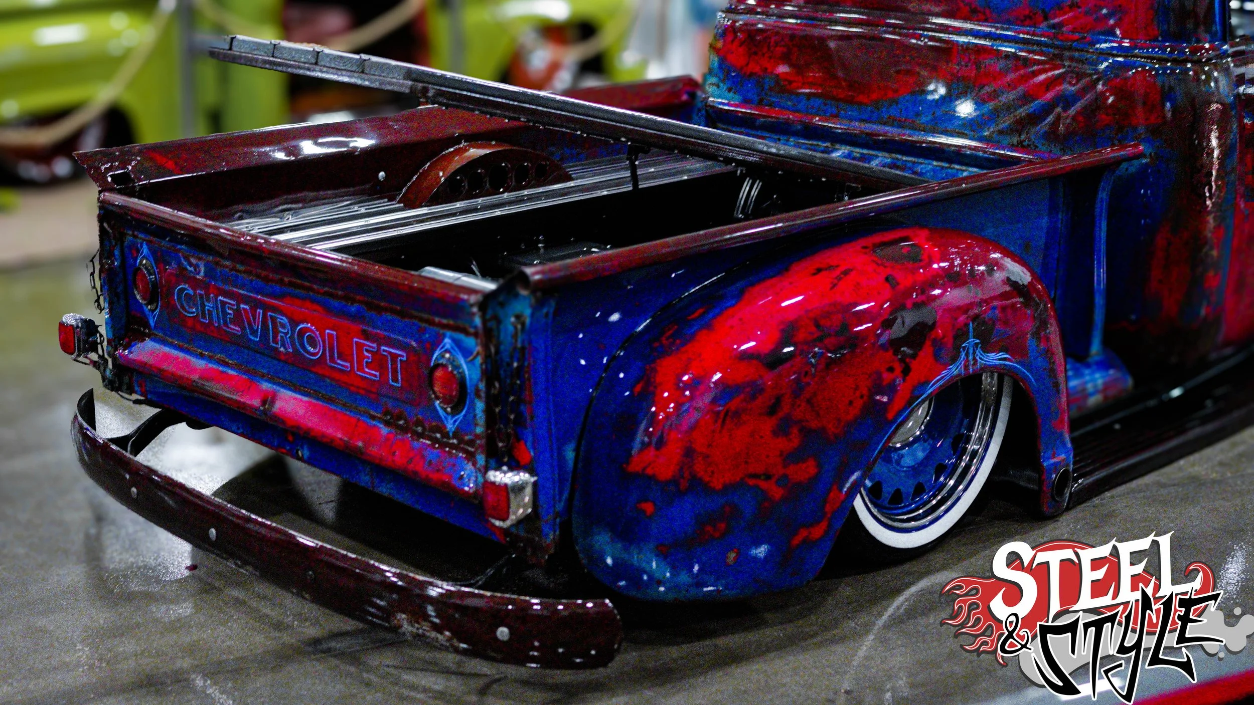 A mini car with a Chevrolet logo, painted in red and blue with a weathered, distressed look, sitting on a wet surface.