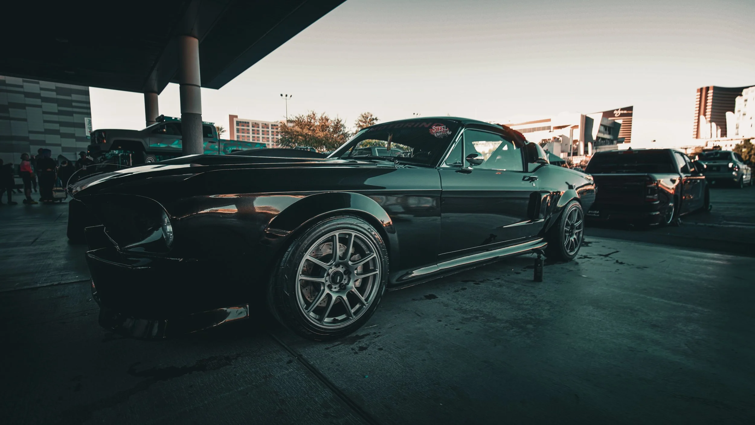 A black vintage muscle car parked at an outdoor car show during sunset, with other vehicles and people in the background.