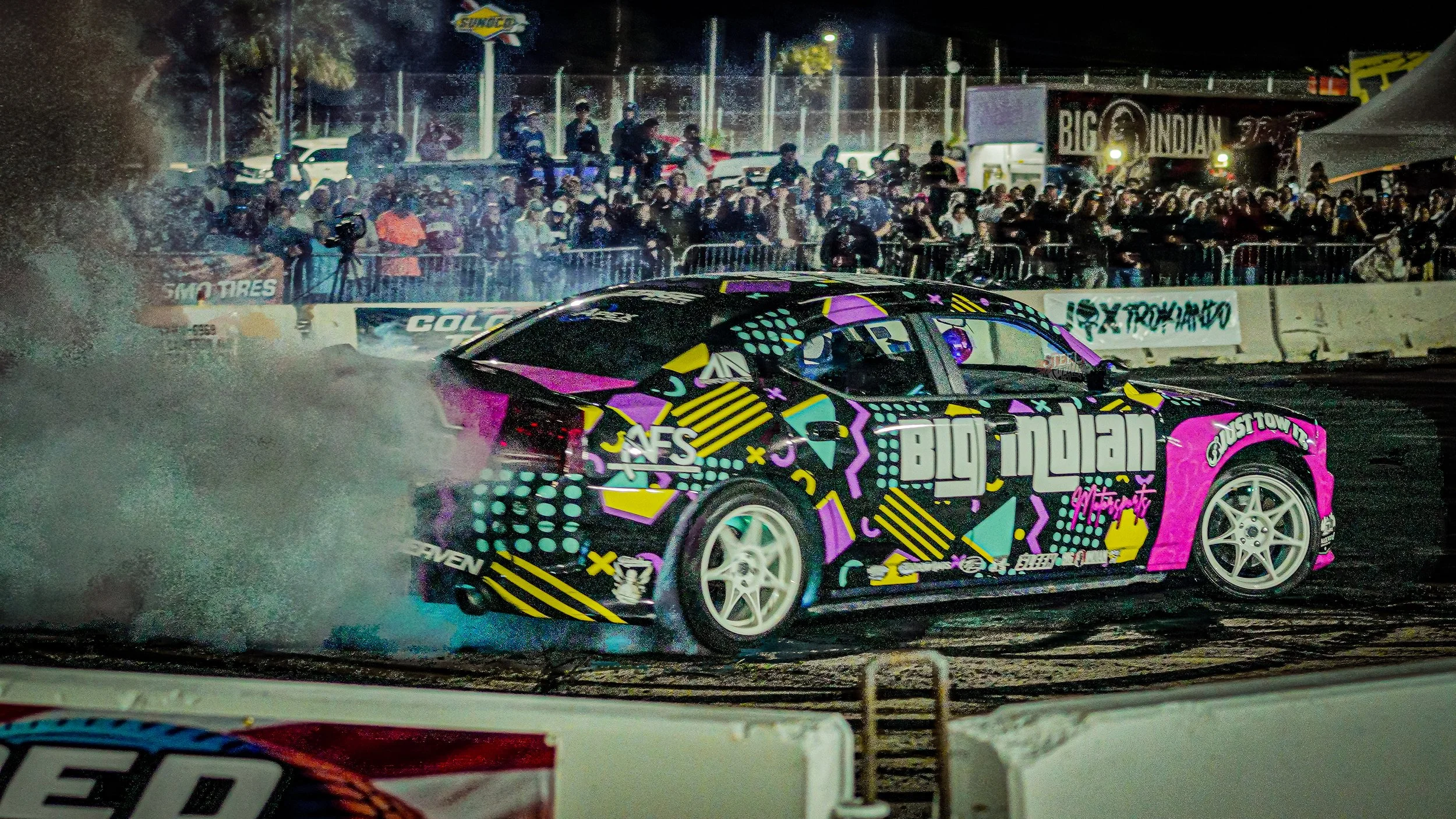 A colorful race car with vibrant geometric graphics and the words 'Big Indian' on its side, performing a burnout at a nighttime motorsport event, creating a cloud of smoke with a large crowd of spectators in the background.