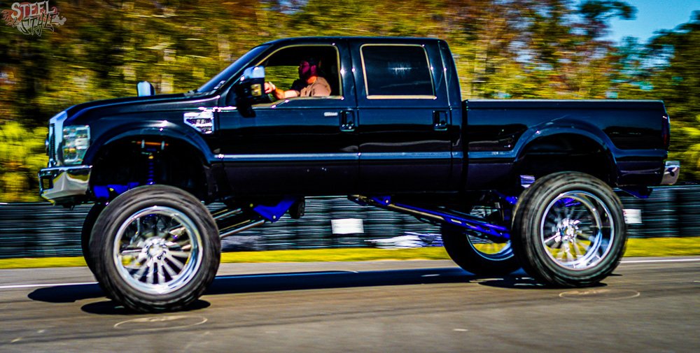 A person driving a black custom lifted truck with large wheels on a race track during daytime.