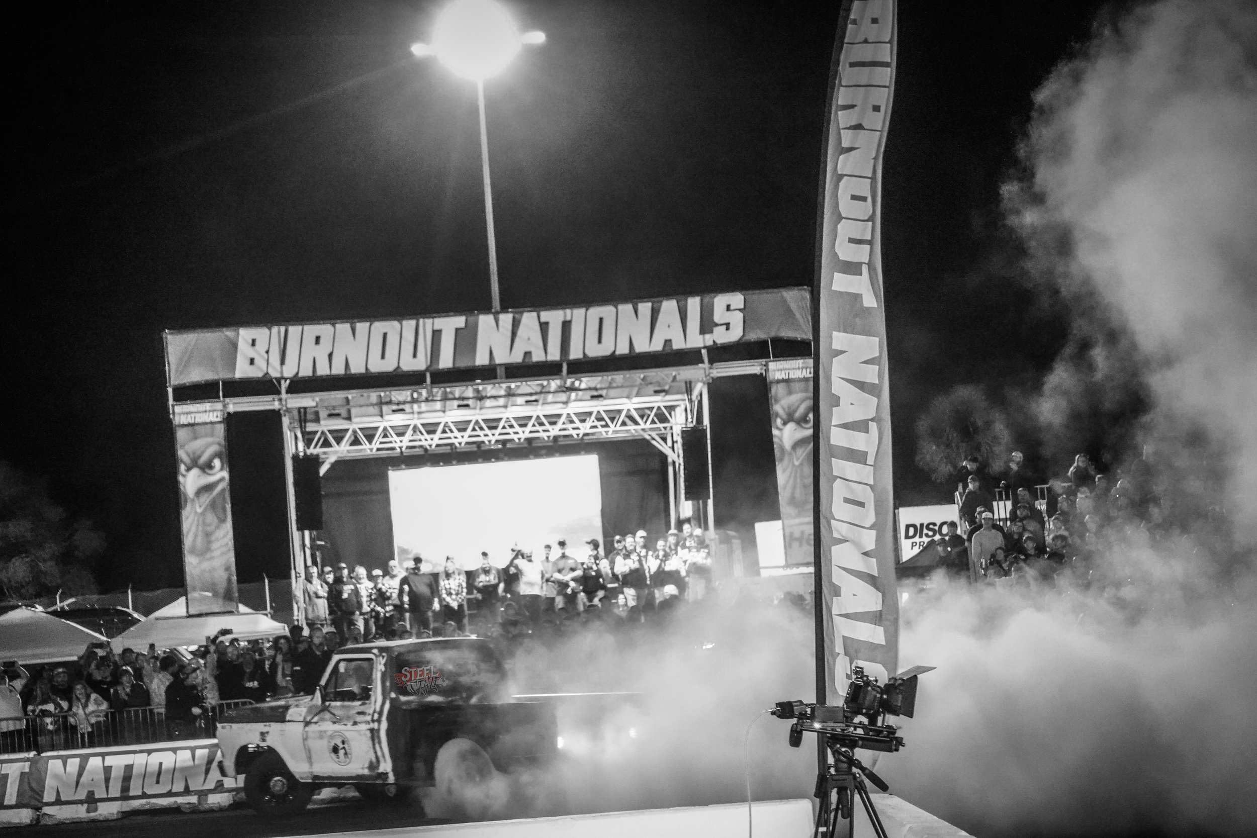 Nighttime at the Burnout Nationals event with a truck performing a burnout, smoke billowing from the tires, and a stage with spectators and participants in the background.