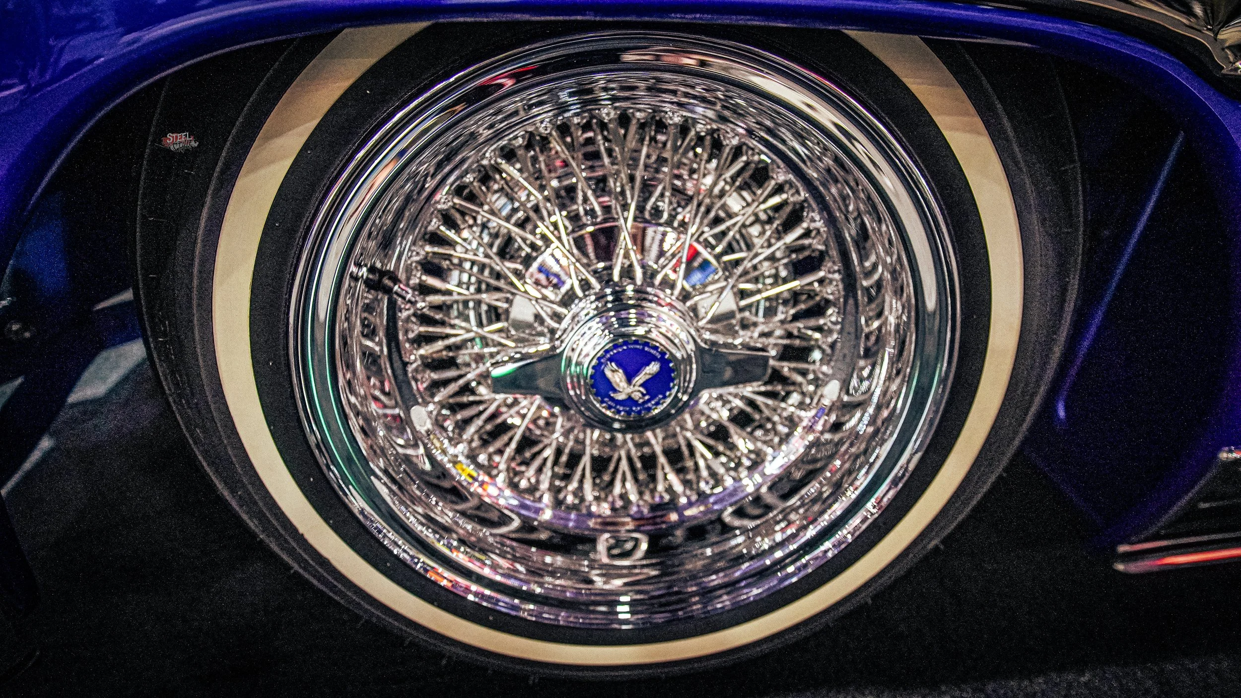 Close-up of a vintage car wheel with a shiny chrome wire-spoke rim and a cream-colored wall tire. The hubcap features a blue emblem with an eagle.