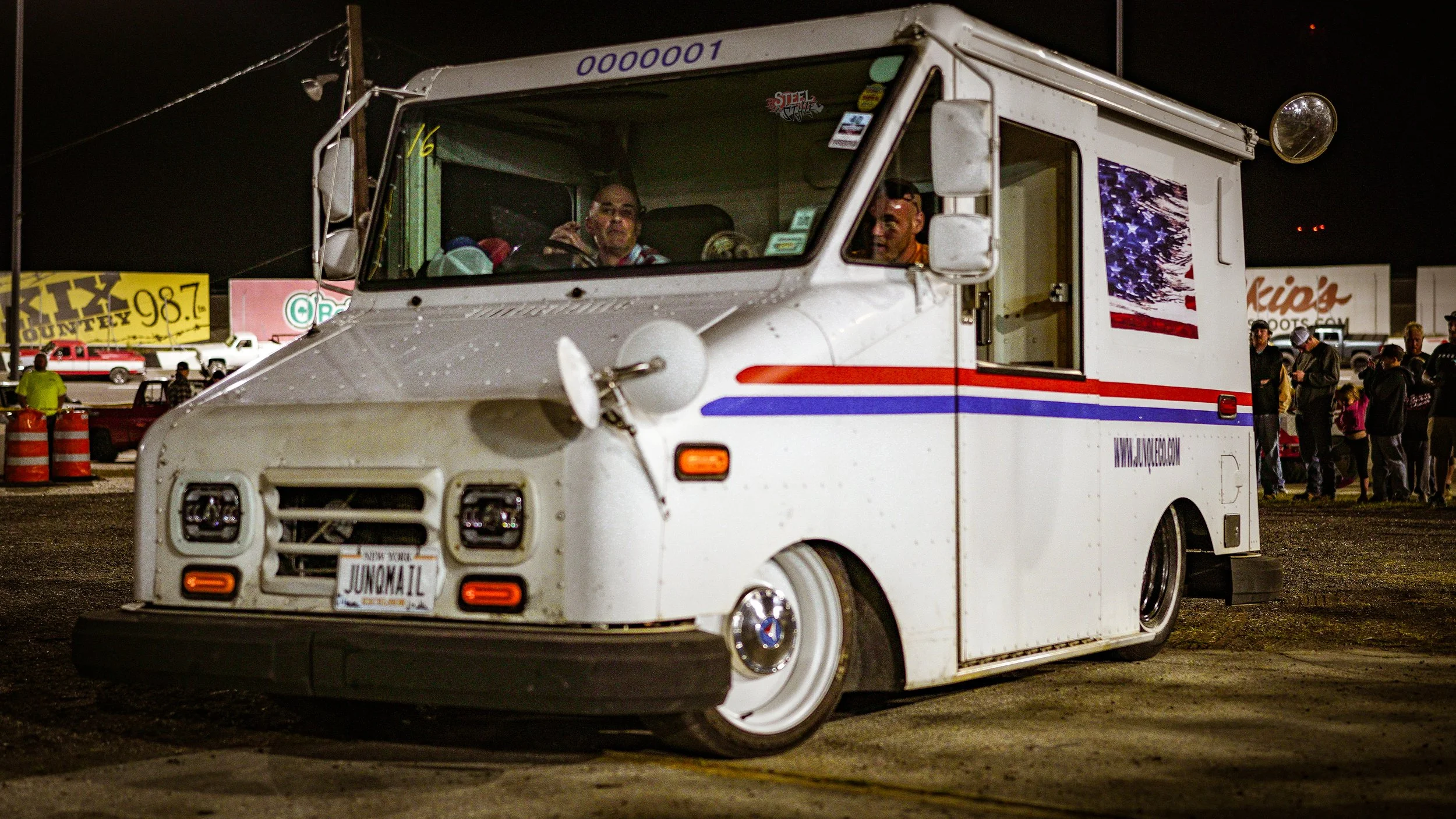 A white ice cream truck with red and blue stripes, a license plate reading 'JUNOMAIL', and an American flag decal on the side, parked at night with a group of people nearby.