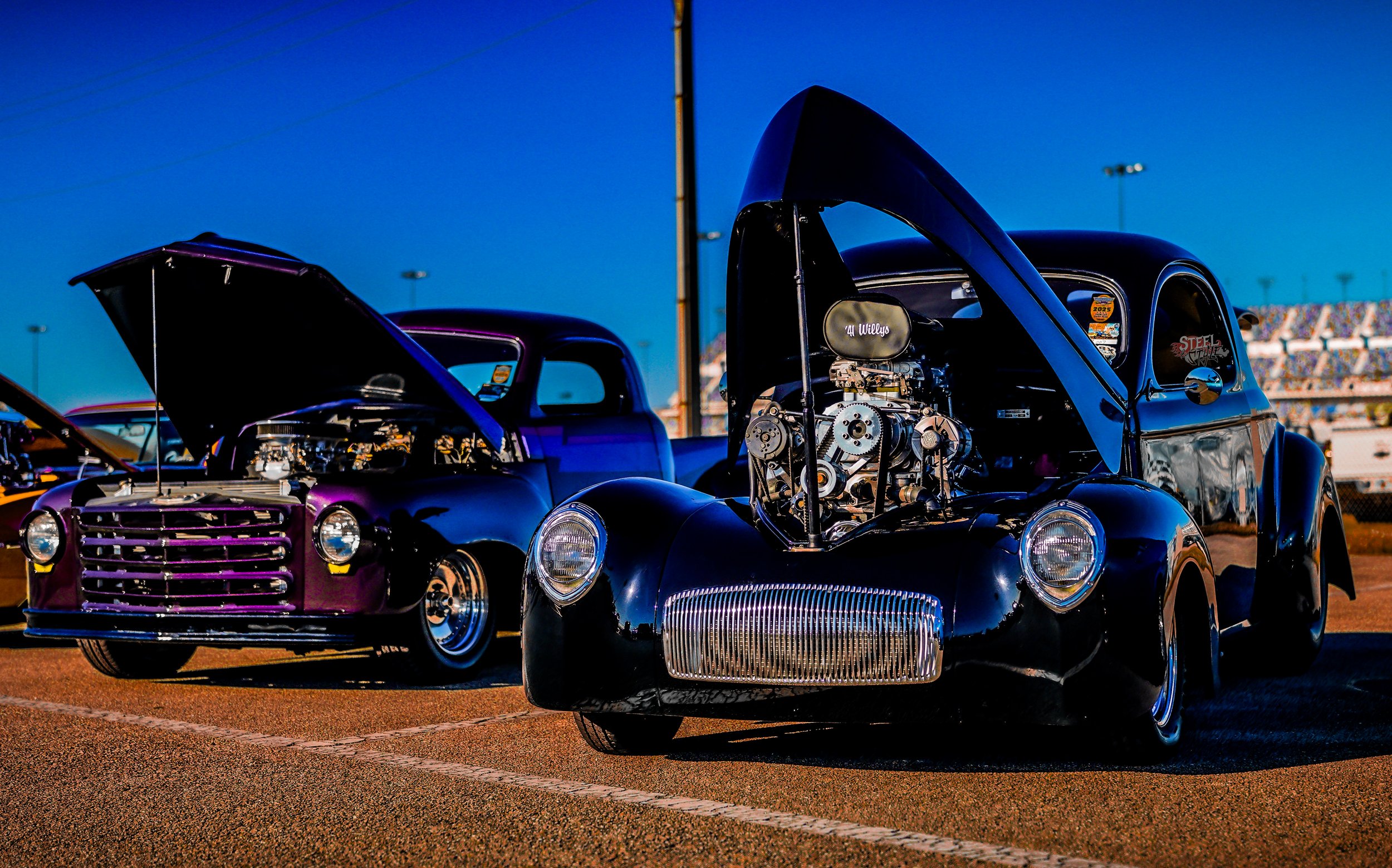 Two vintage drag racing cars with open hoods, parked on a race track during sunset, showcasing their engines.