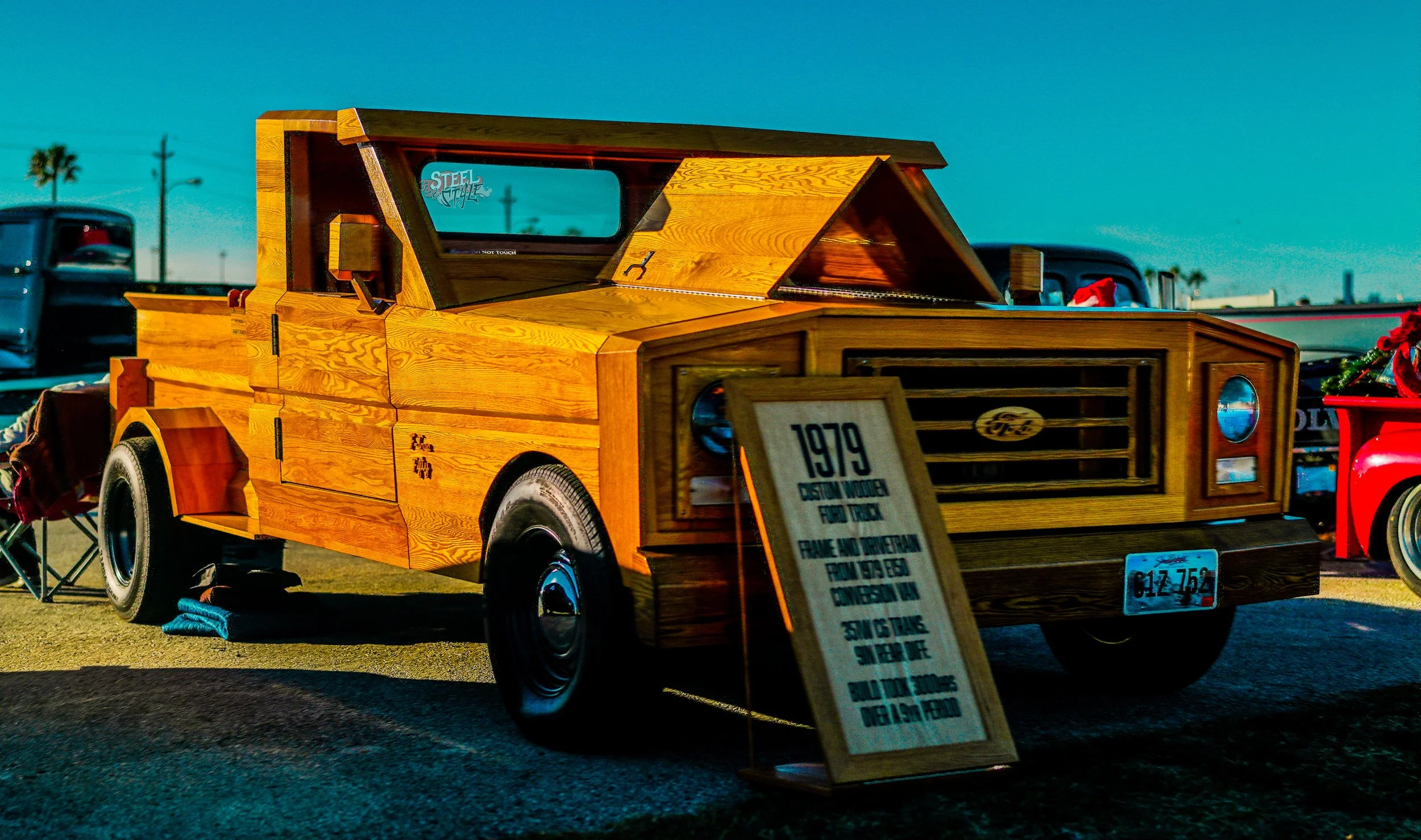 A vintage truck designed to look like it's made of wood, on display at a car show, with a sign in front detailing its history and features.