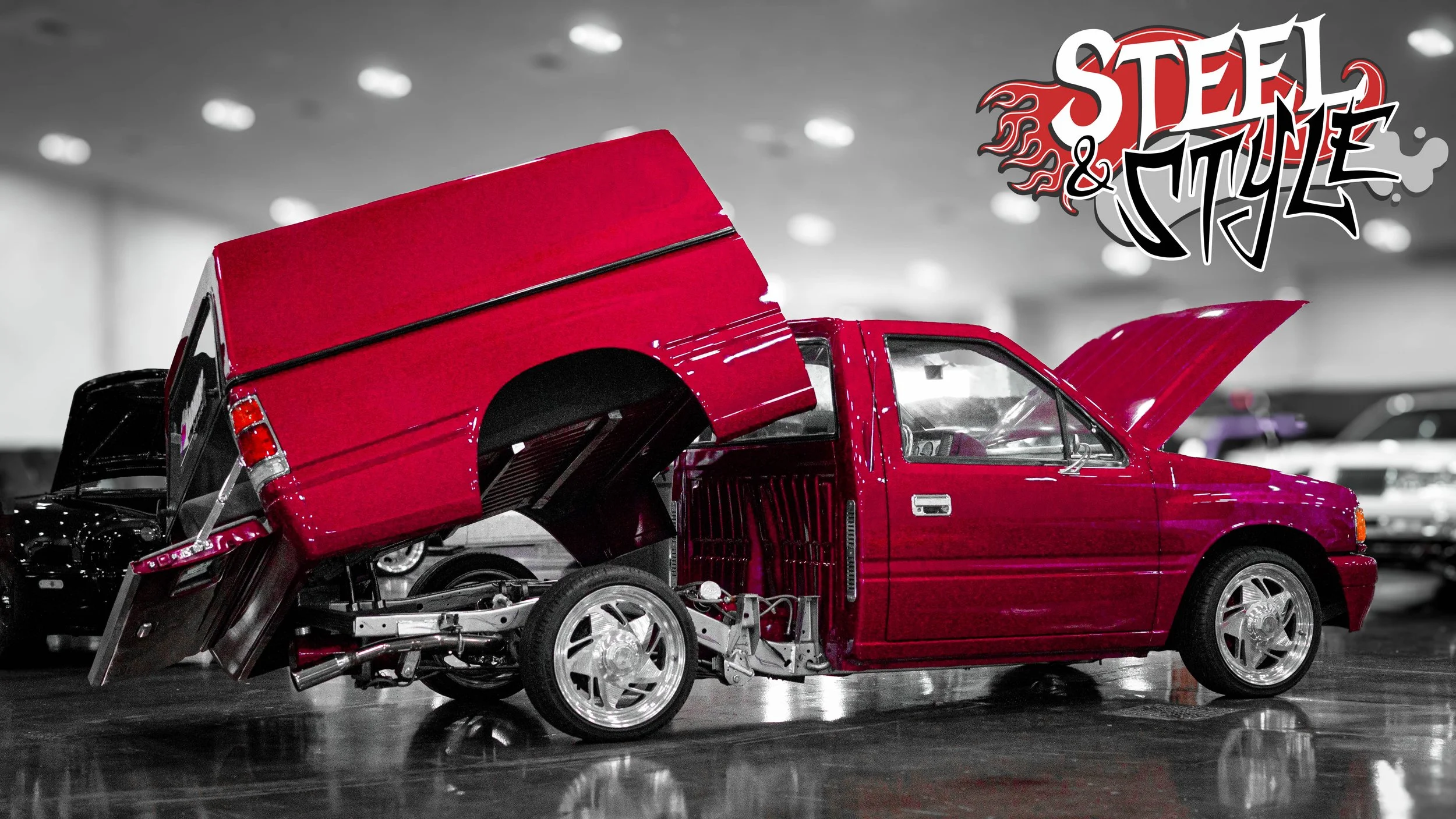 A red modified car with its hood open and a raised rear section, displayed indoors with a gray background and other vehicles.