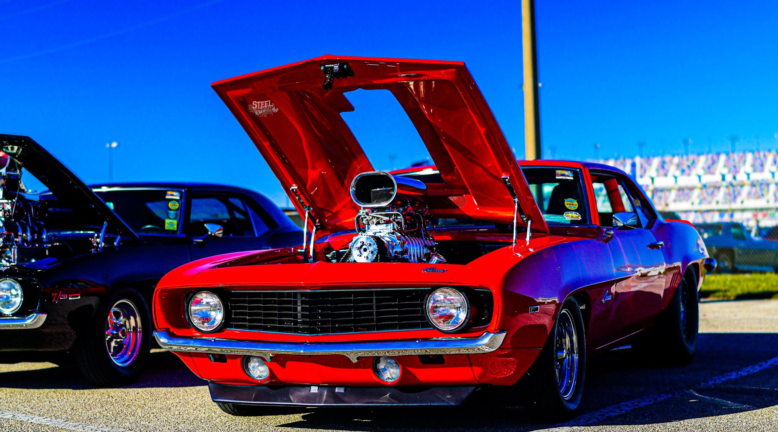 Red vintage muscle car with an open hood displaying its engine, parked among other classic cars at a car show.
