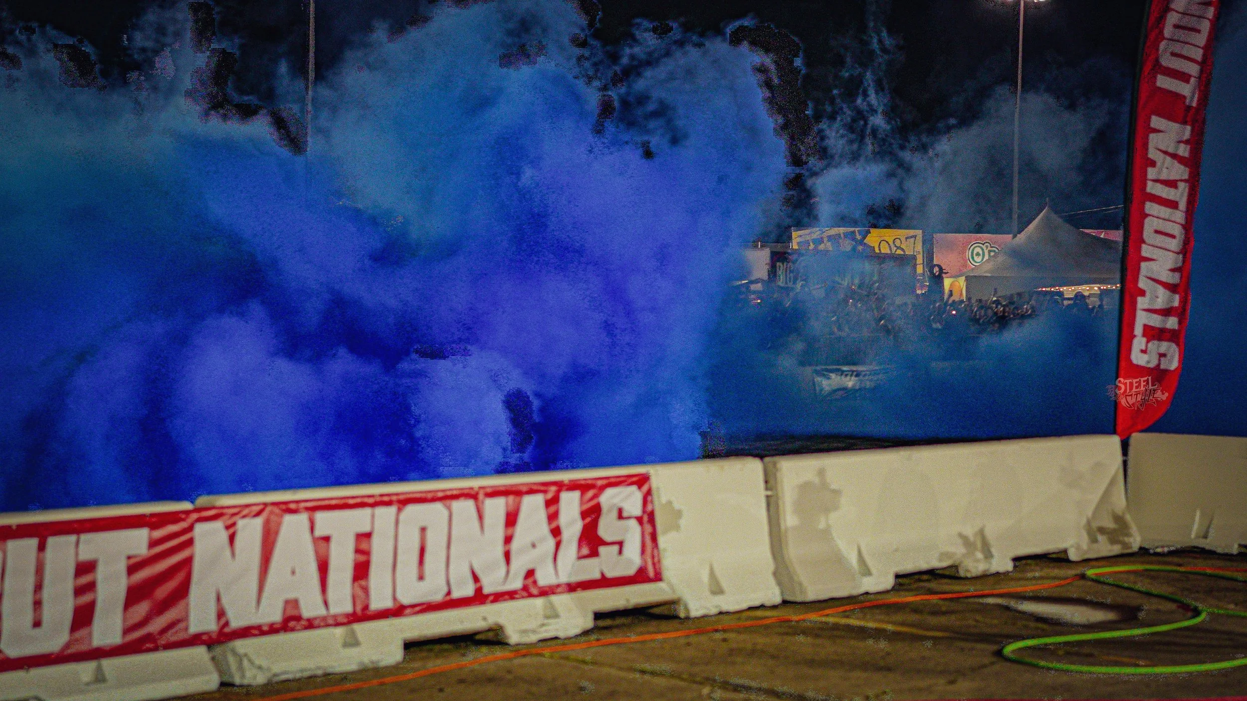 Scene at a racing event with blue smoke in the air, barriers with 'NATIONALS' written on them, tents, and people in the background.