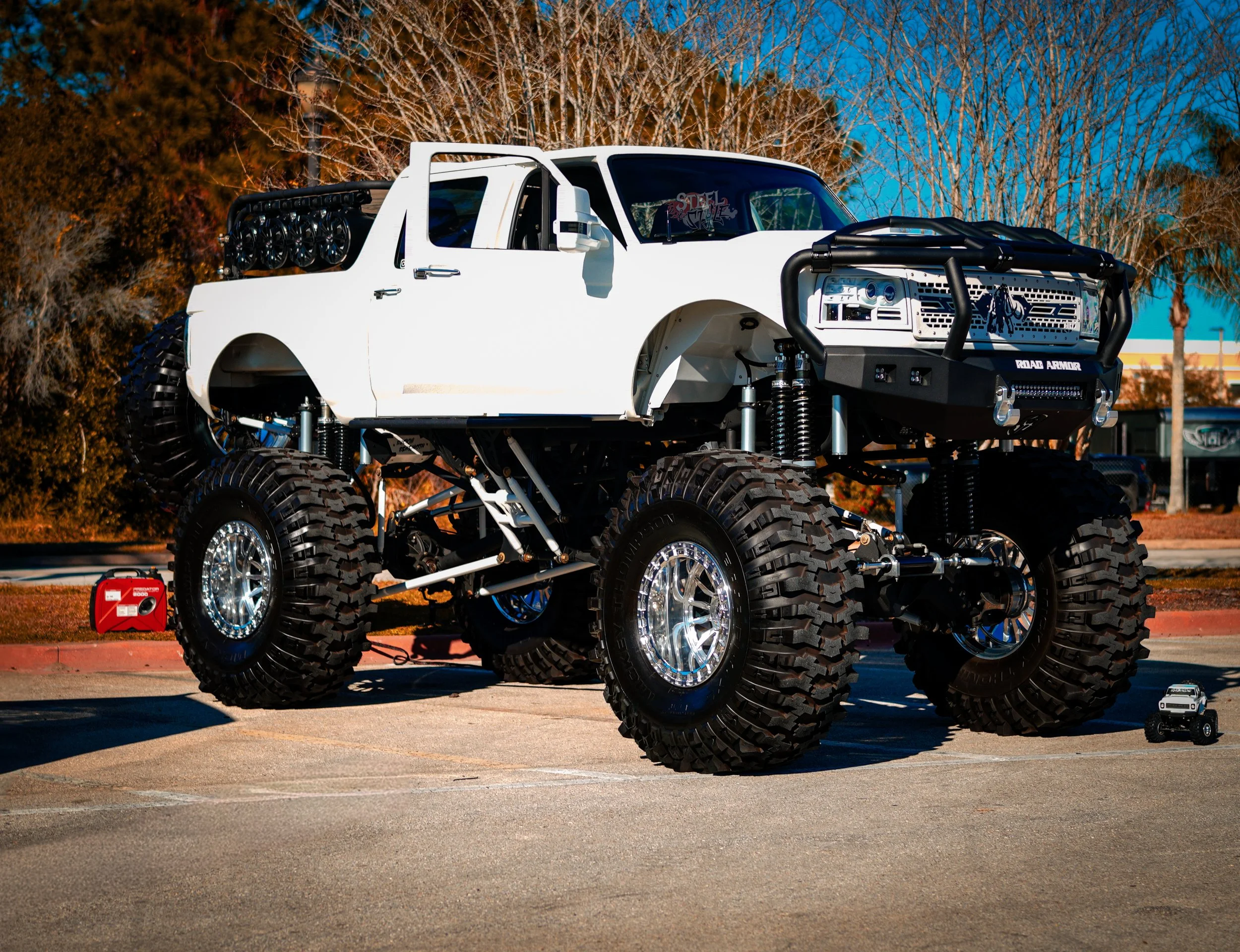 A heavily modified off-road truck with large tires, a white body, and a front bull bar, parked in a lot with leafless trees and a blue sky in the background.