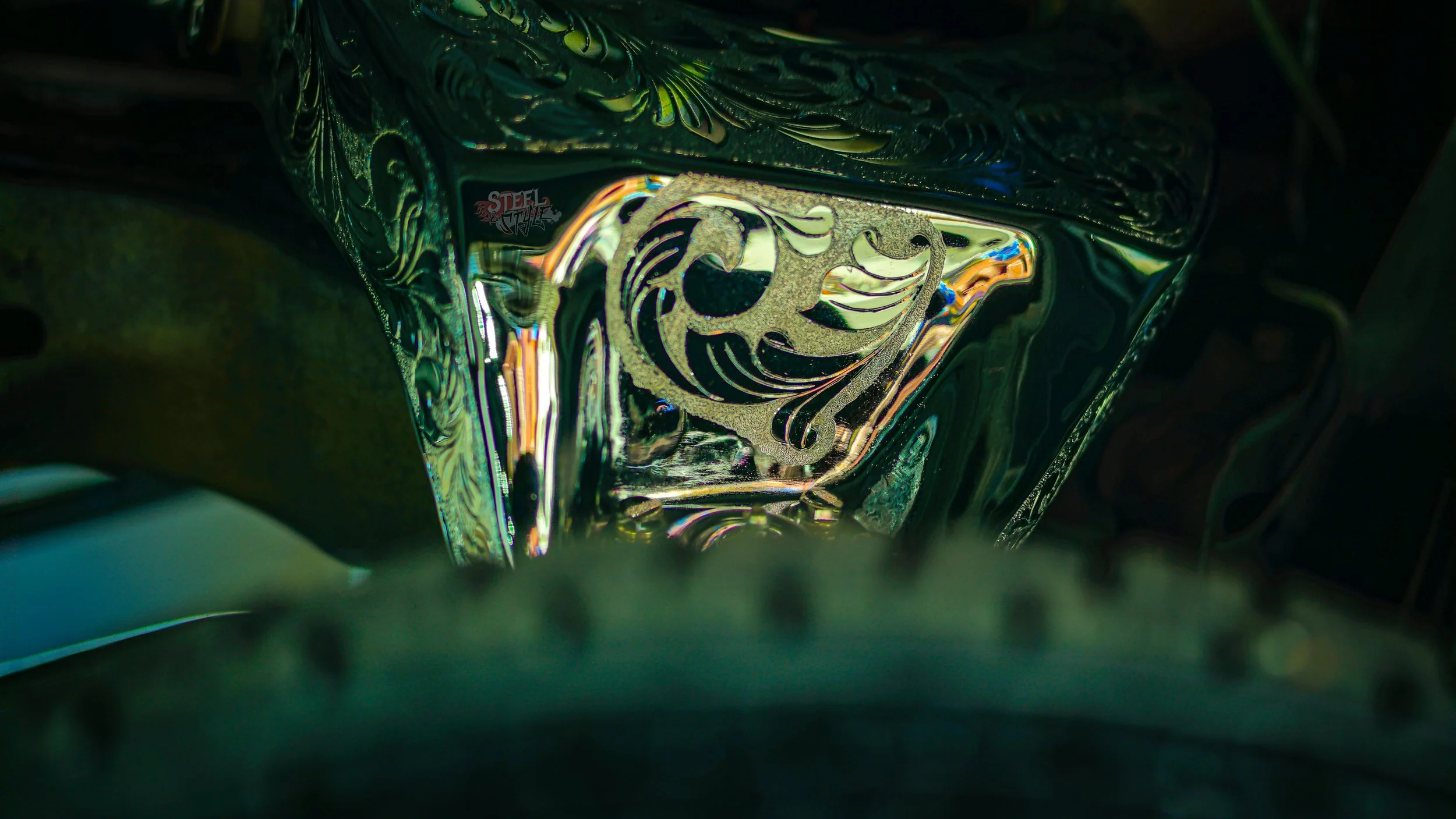 A close-up photograph of an intricately designed, reflective metallic object, possibly a part of machinery or a decorative art piece, with swirling and floral patterns.