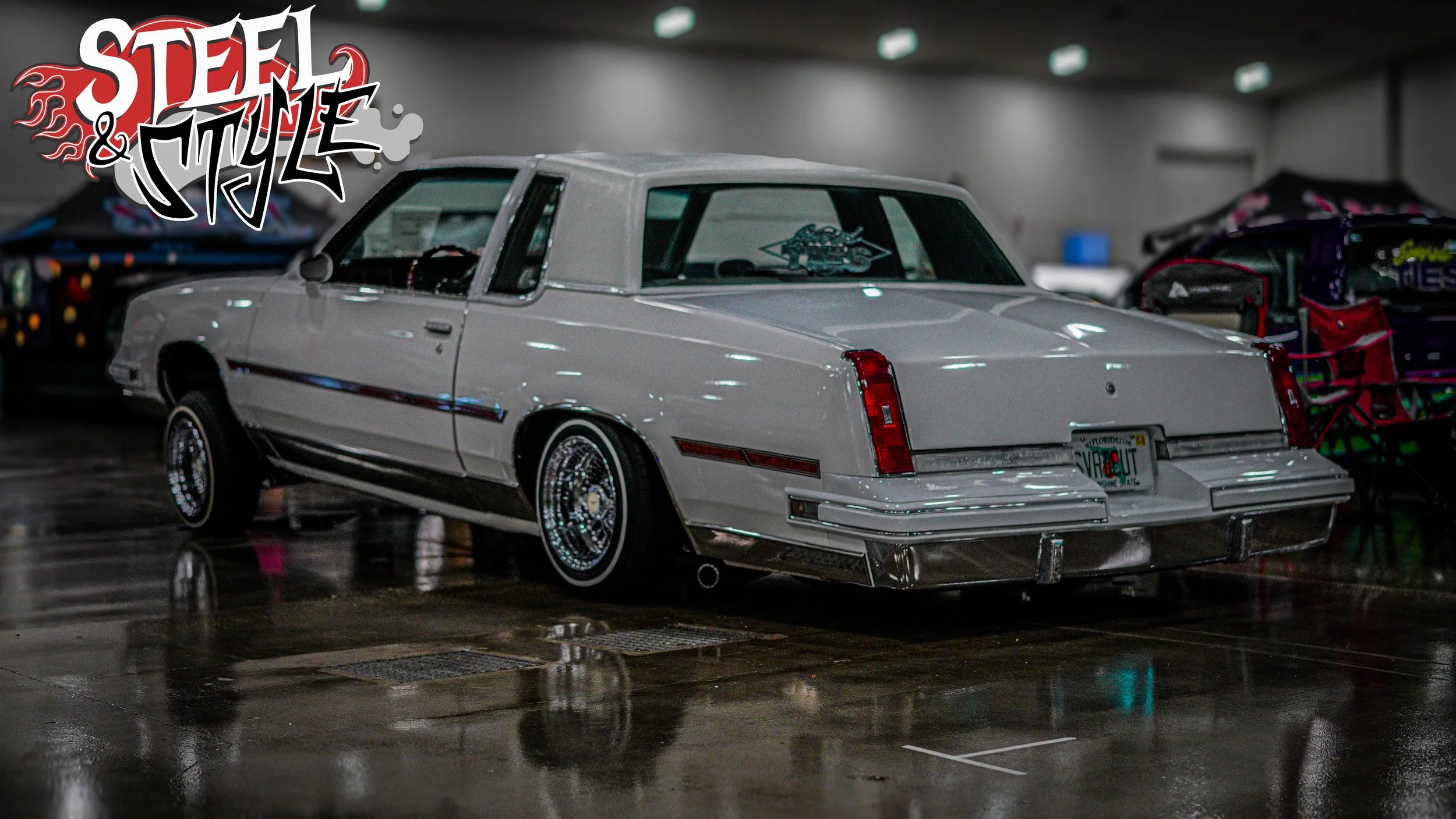 A classic white Cadillac with a vinyl roof is parked indoors at an exhibition, with a 'Steel & Style' logo in the top left corner and other cars in the background.