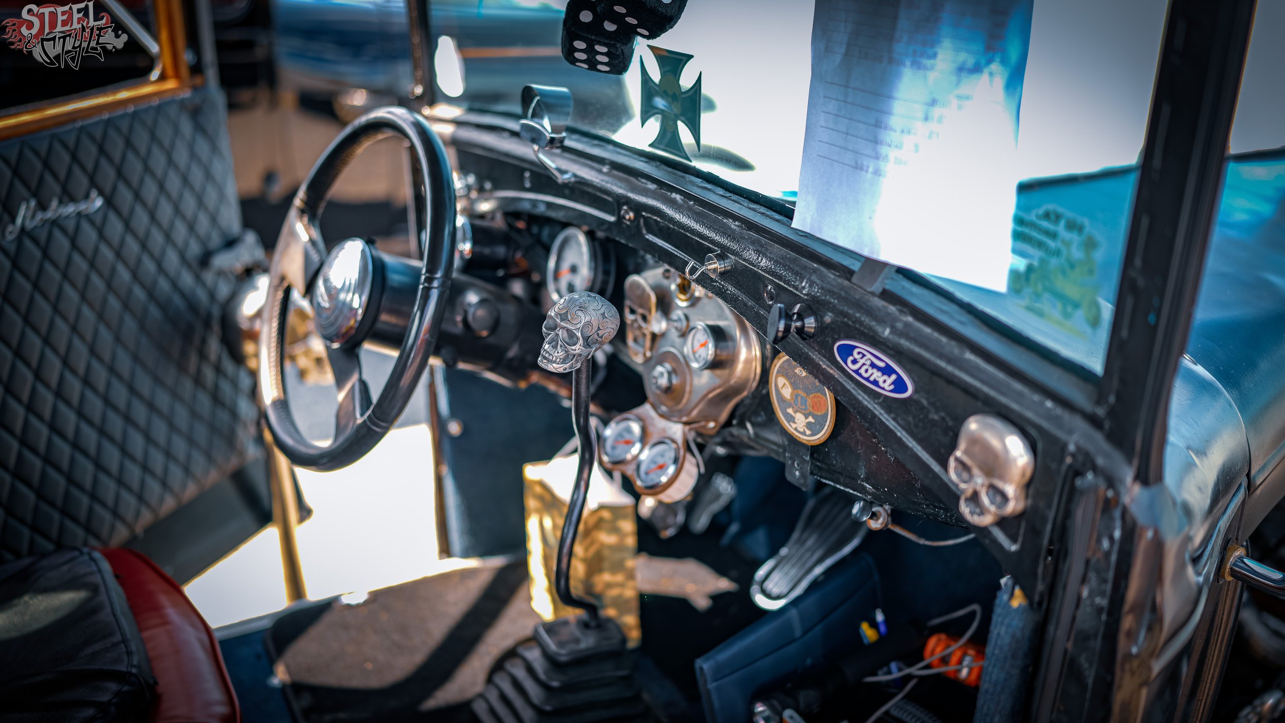 Close-up of a vintage car's interior, featuring a skull gear shift knob, a steering wheel, and a dashboard with various gauges. A mouse decal is hanging on the rearview mirror, and there are stickers on the dashboard, including a Ford logo.