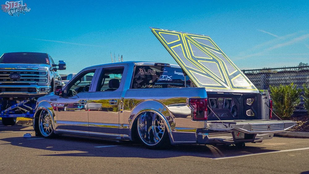 A highly customized silver pickup truck with a reflective chrome finish, fitted with large wheels and low-profile tires, parked outdoors on a sunny day. The truck has an open tailgate revealing a modified bed with a large, illuminated geometric-style