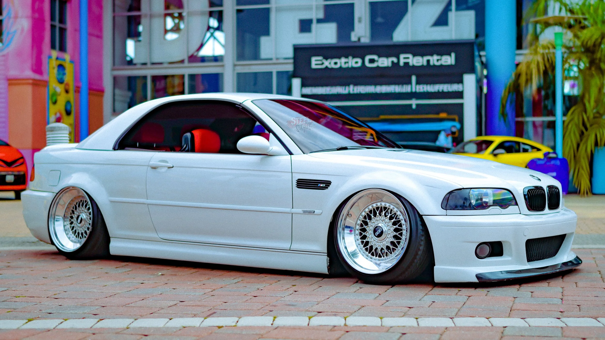 A white modified BMW car with lowered suspension and custom multi-spoke rims parked on a brick sidewalk in front of a colorful amusement park.