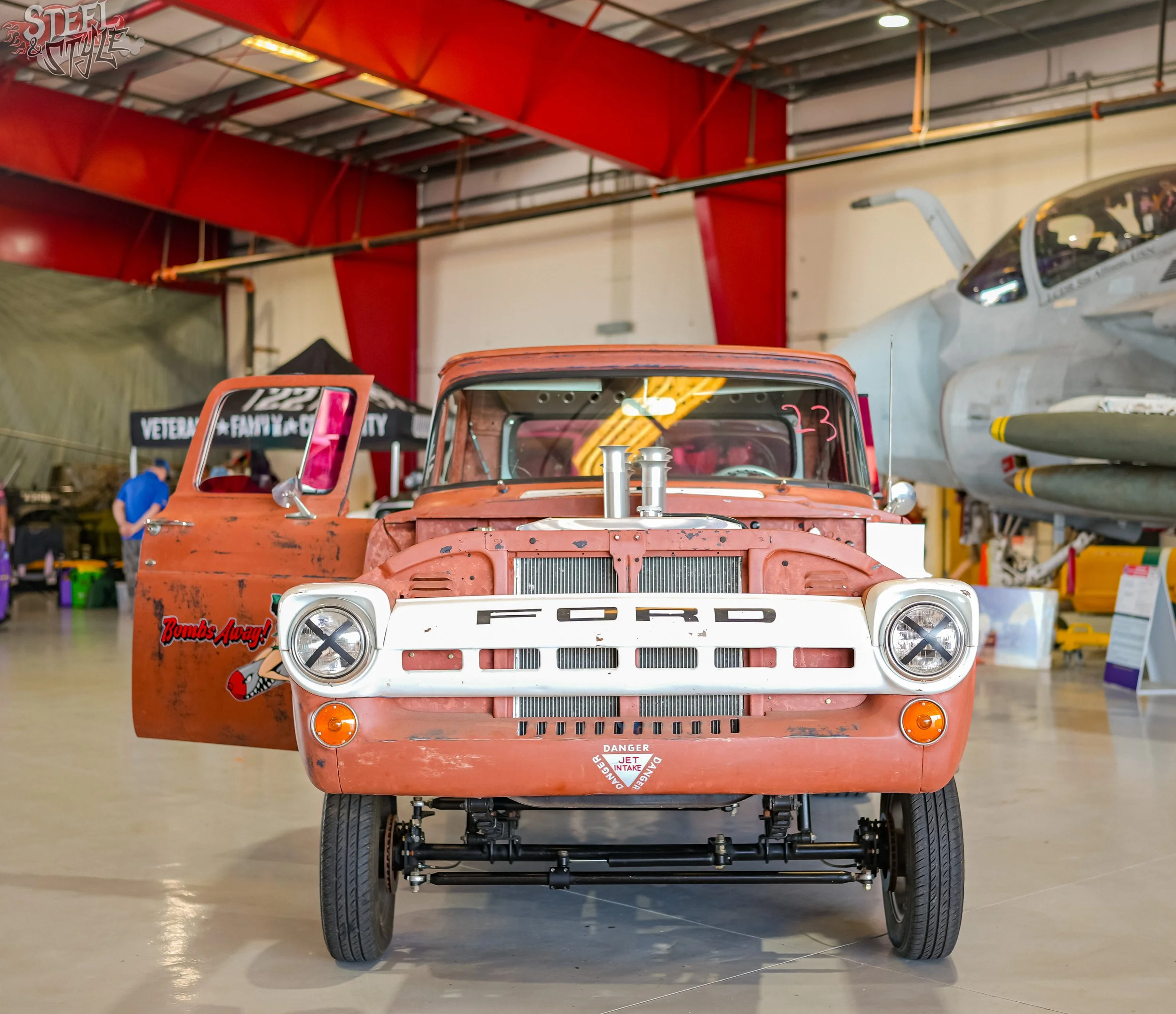 Front view of a vintage orange truck in an indoor aviation or automotive museum, with airplane models and display booths visible in the background.