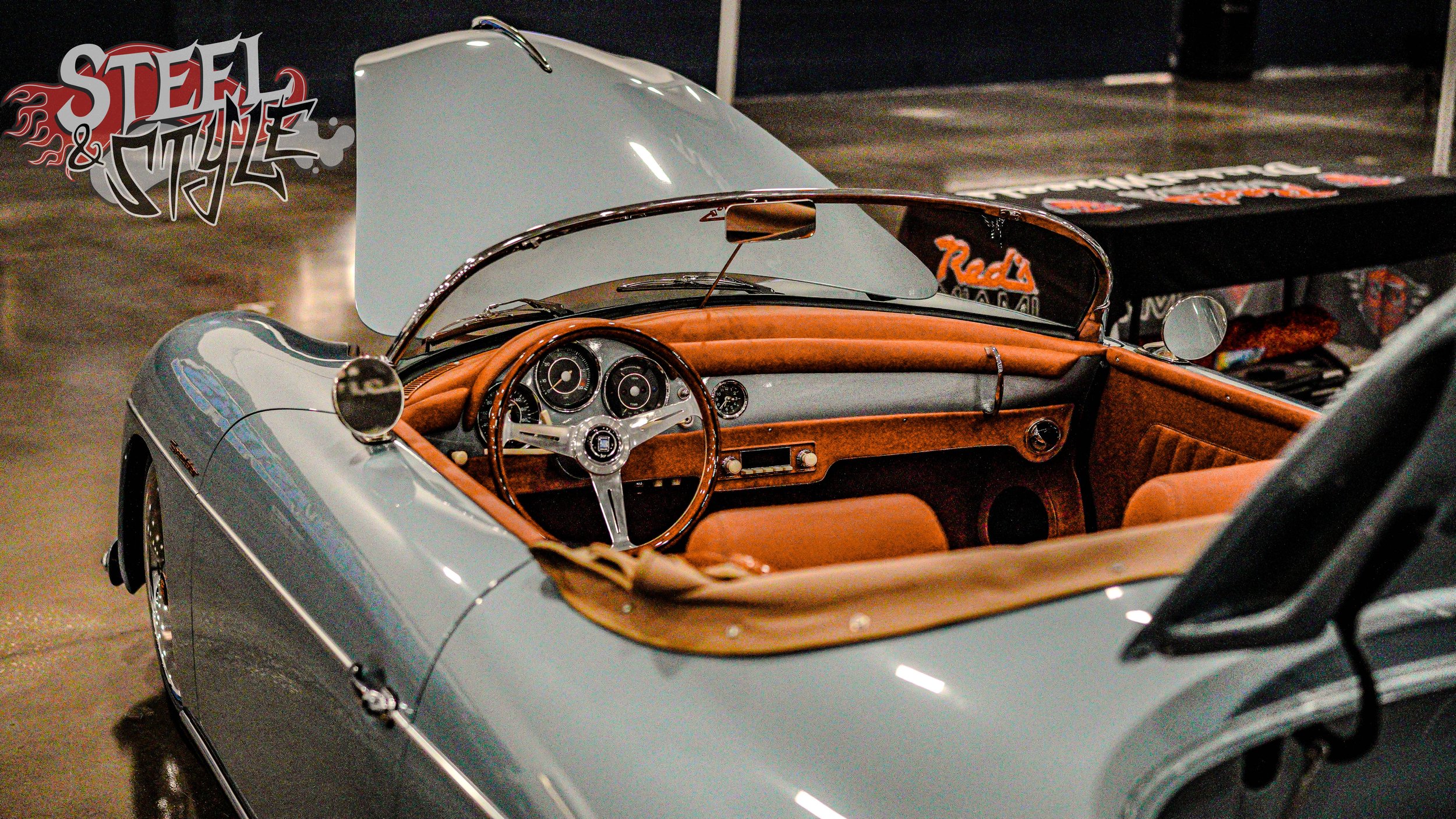 A vintage convertible sports car with a light blue exterior, brown leather interior, and a classic dashboard with analog gauges, displayed at a car exhibition.