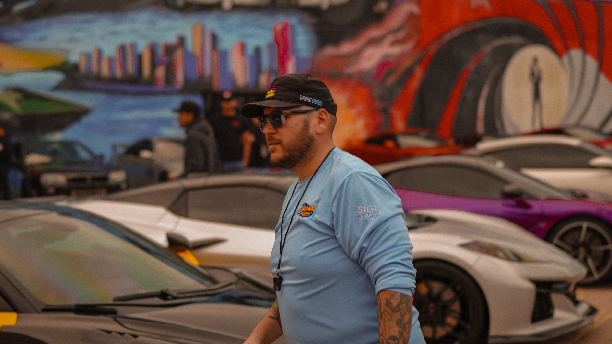 A man wearing sunglasses, a cap, and a blue shirt standing among luxury sports cars at a car show with colorful graffiti art in the background.