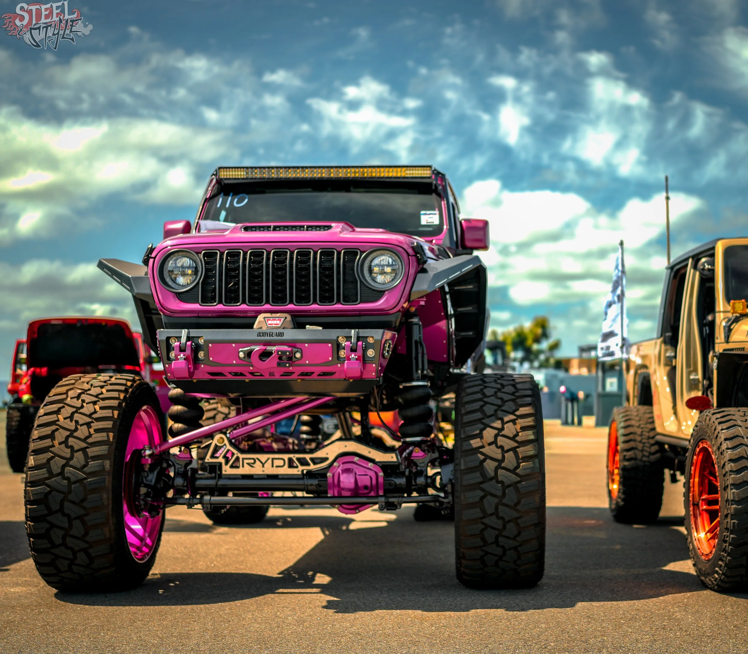 A pink off-road vehicle with large tires, a prominent front grille, and a light bar on top, parked at an outdoor event under a cloudy sky.