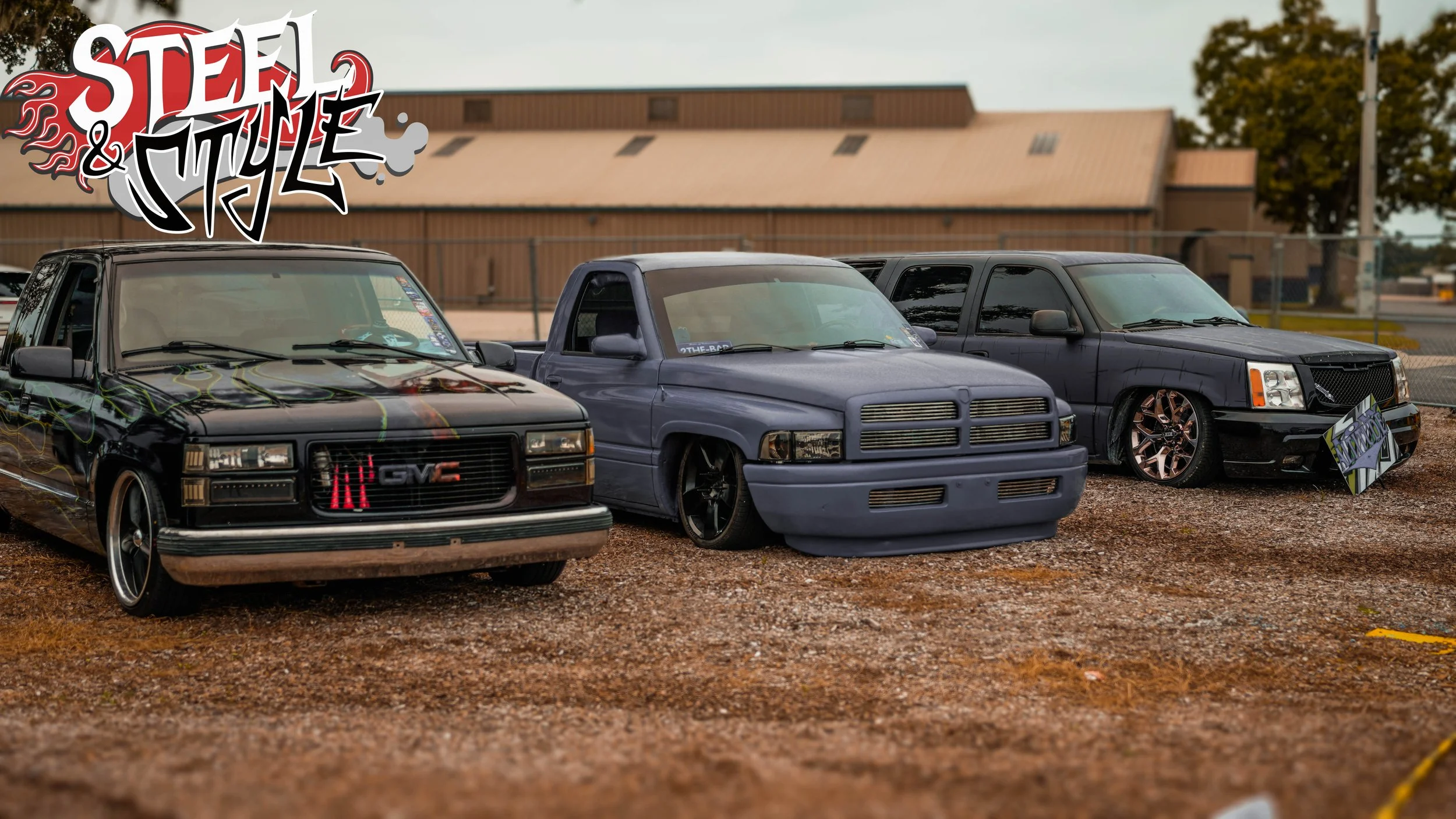 Three customized pickup trucks parked on a gravel lot with a building and trees in the background. The left truck has a black paint job with graffiti-style design, the middle truck is painted in matte blue with a lowered stance, and the right truck i