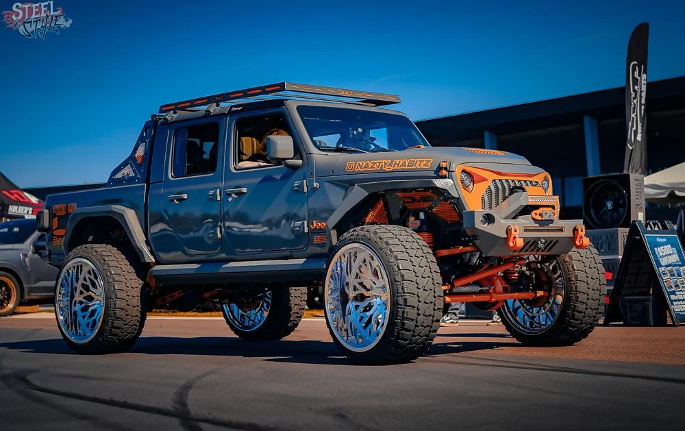 Modified Jeep truck with large off-road tires, custom wheels, and orange accents at a car show.
