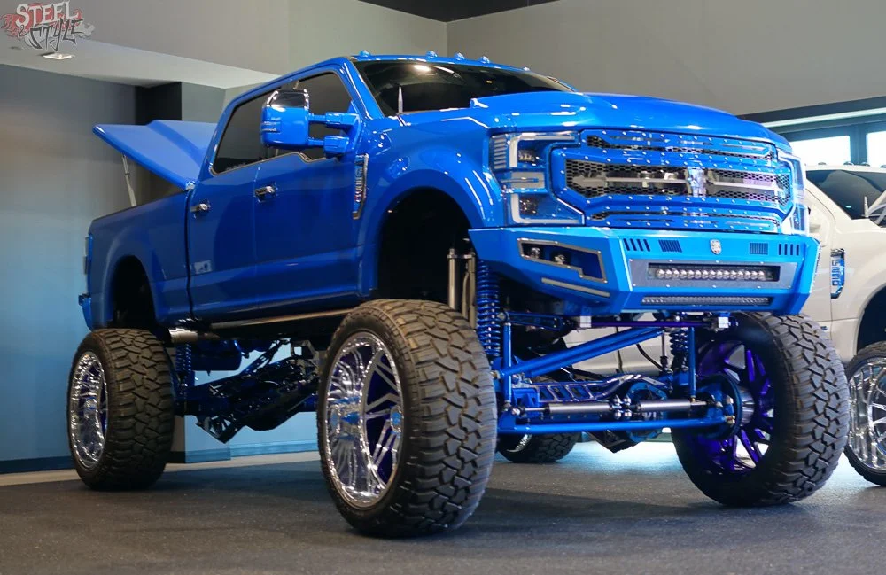 A custom blue lifted pickup truck with large off-road tires and chrome rims, displayed indoors at an exhibition.