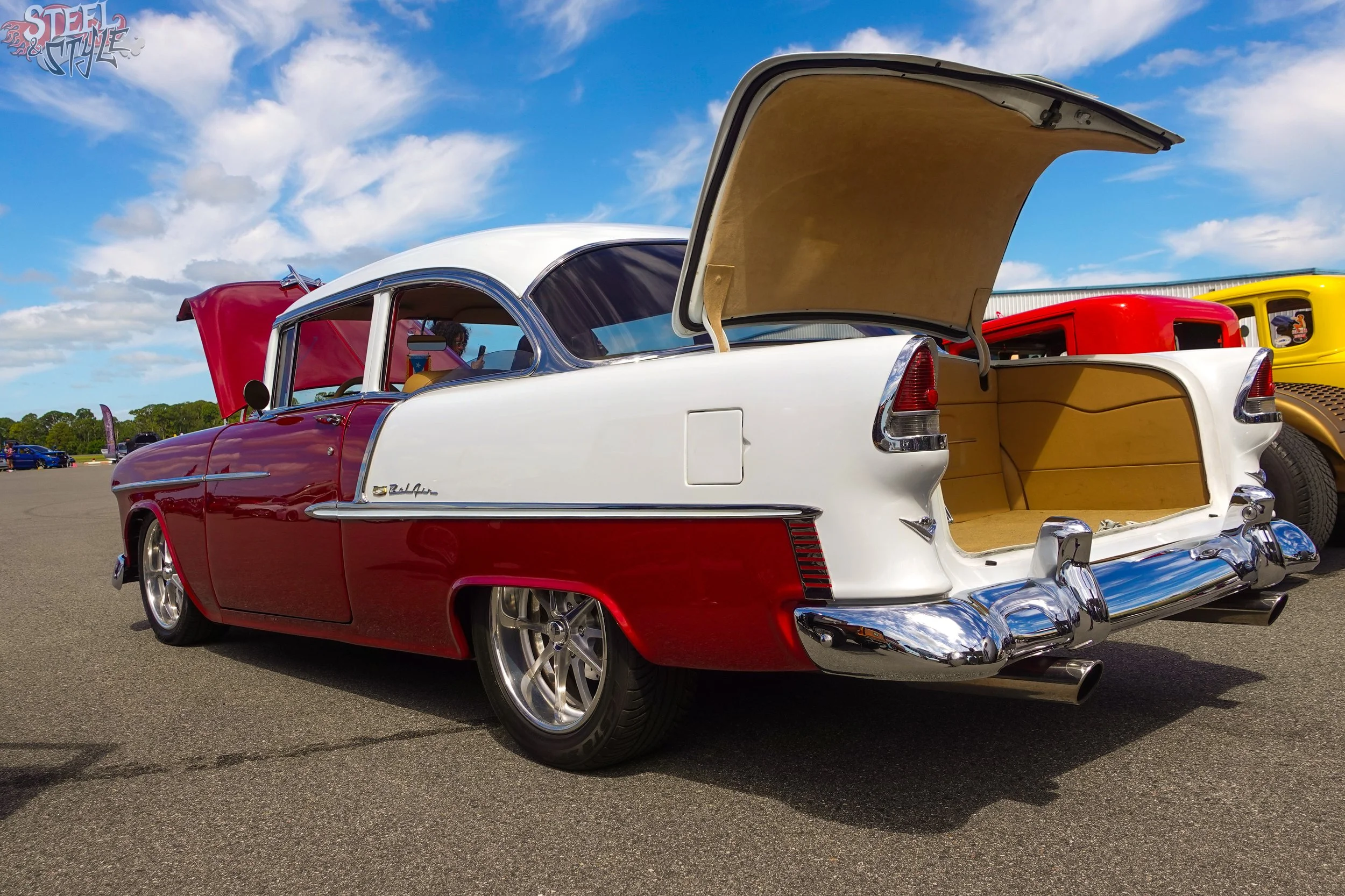 A classic white and red vintage car with open trunk displaying a beige interior at an outdoor car show under a partly cloudy sky.