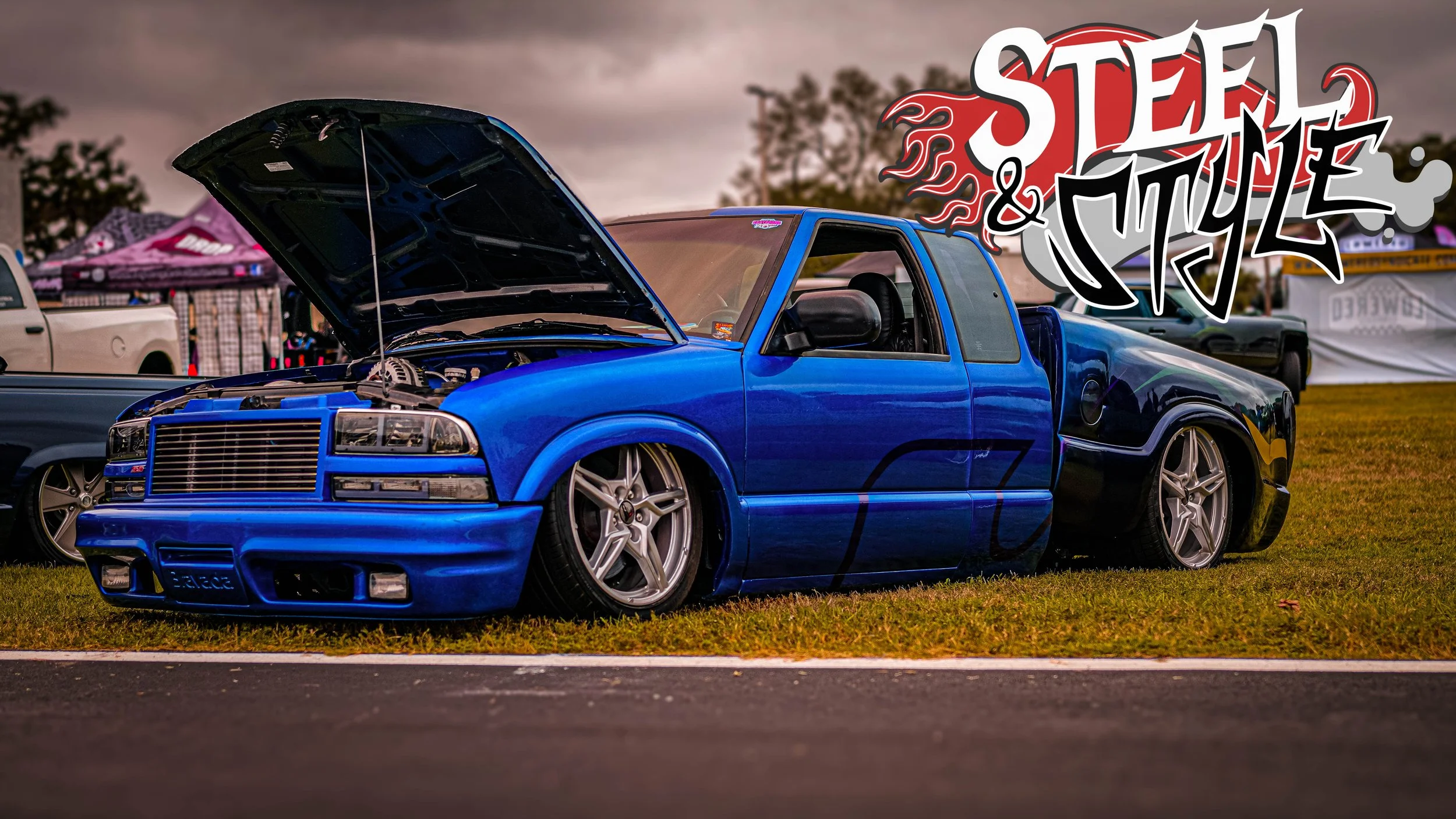 Blue modified car with open hood at a car show, with a custom graphic 'Steel & Style' above it.