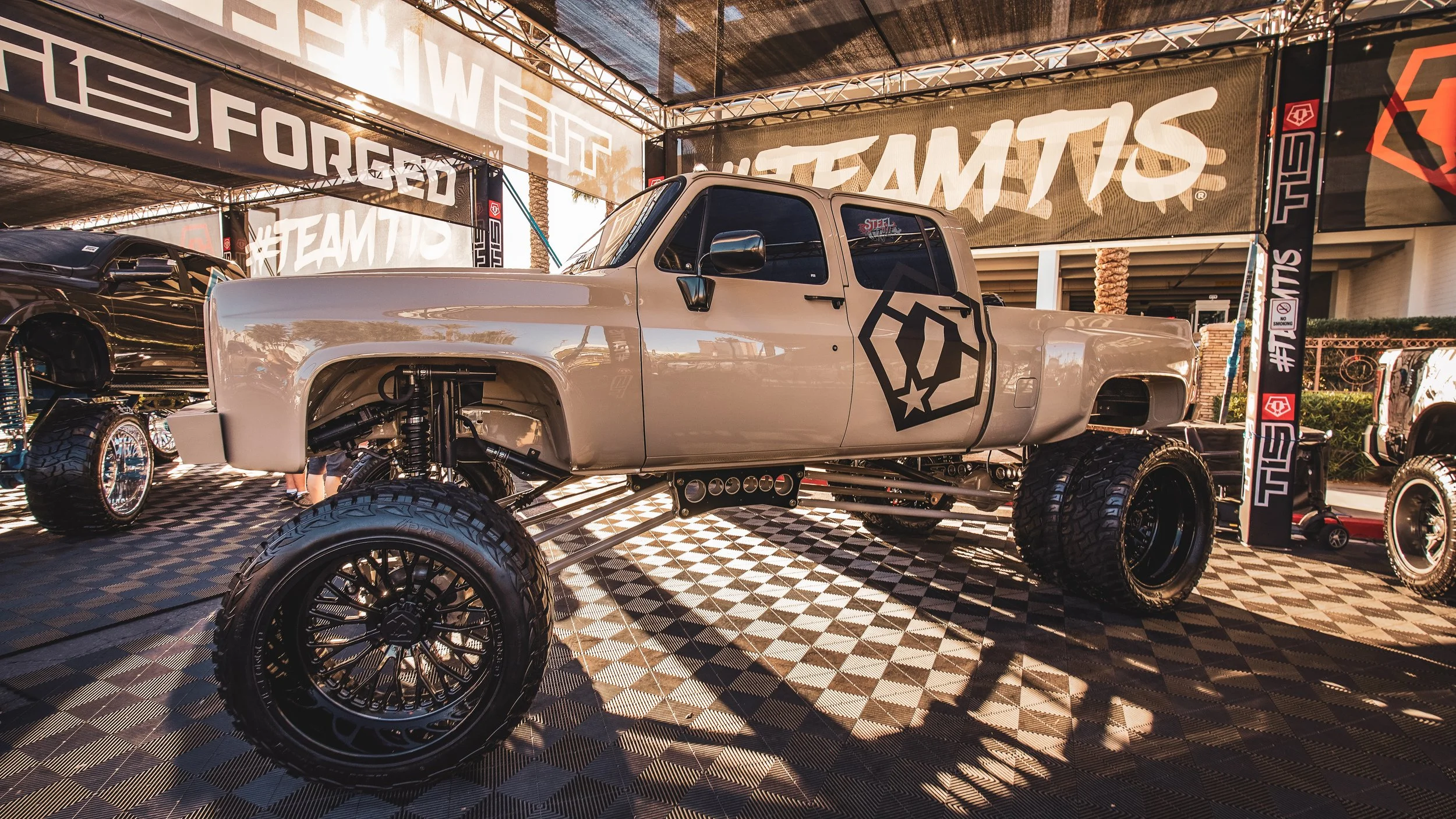 Customized off-road pickup truck with lifted suspension and large tires on display at a promotional event, with banners and signage in the background.