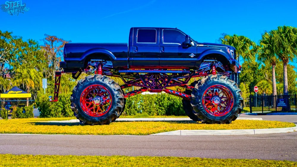 A giant monster truck with oversized tires and a black truck cab, prominently displayed on a grassy area surrounded by trees and a road.