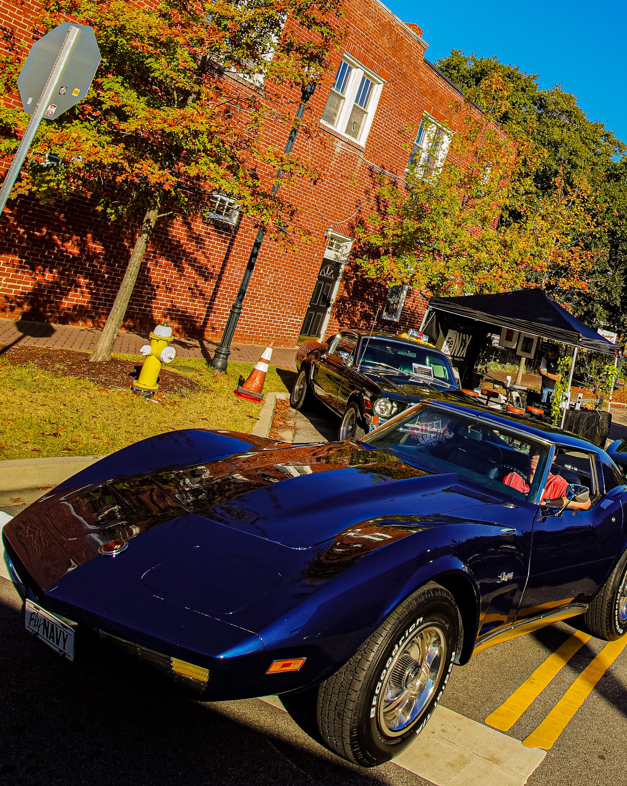 A vintage blue Corvette parked on a city street during a car show, with other classic cars visible in the background, under trees with fall foliage and a brick building.