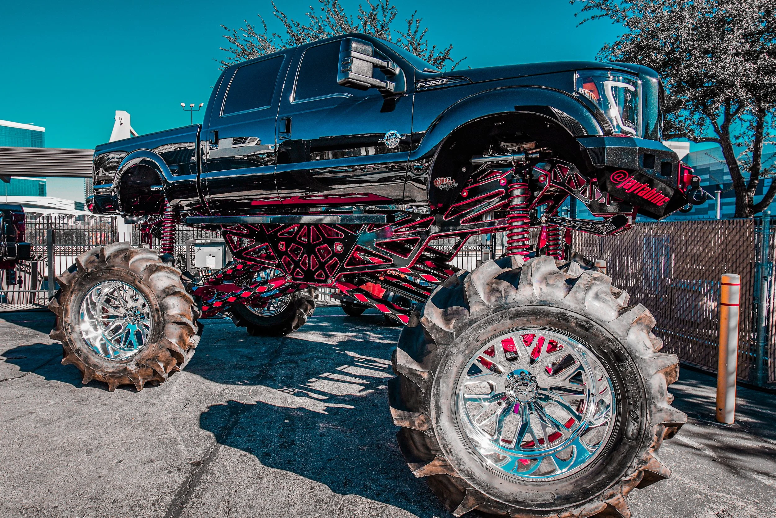 A monster truck with a lifted black pickup body, large tires, and a pink-trimmed suspension system, parked outdoors in a lot with a fence and trees in the background.