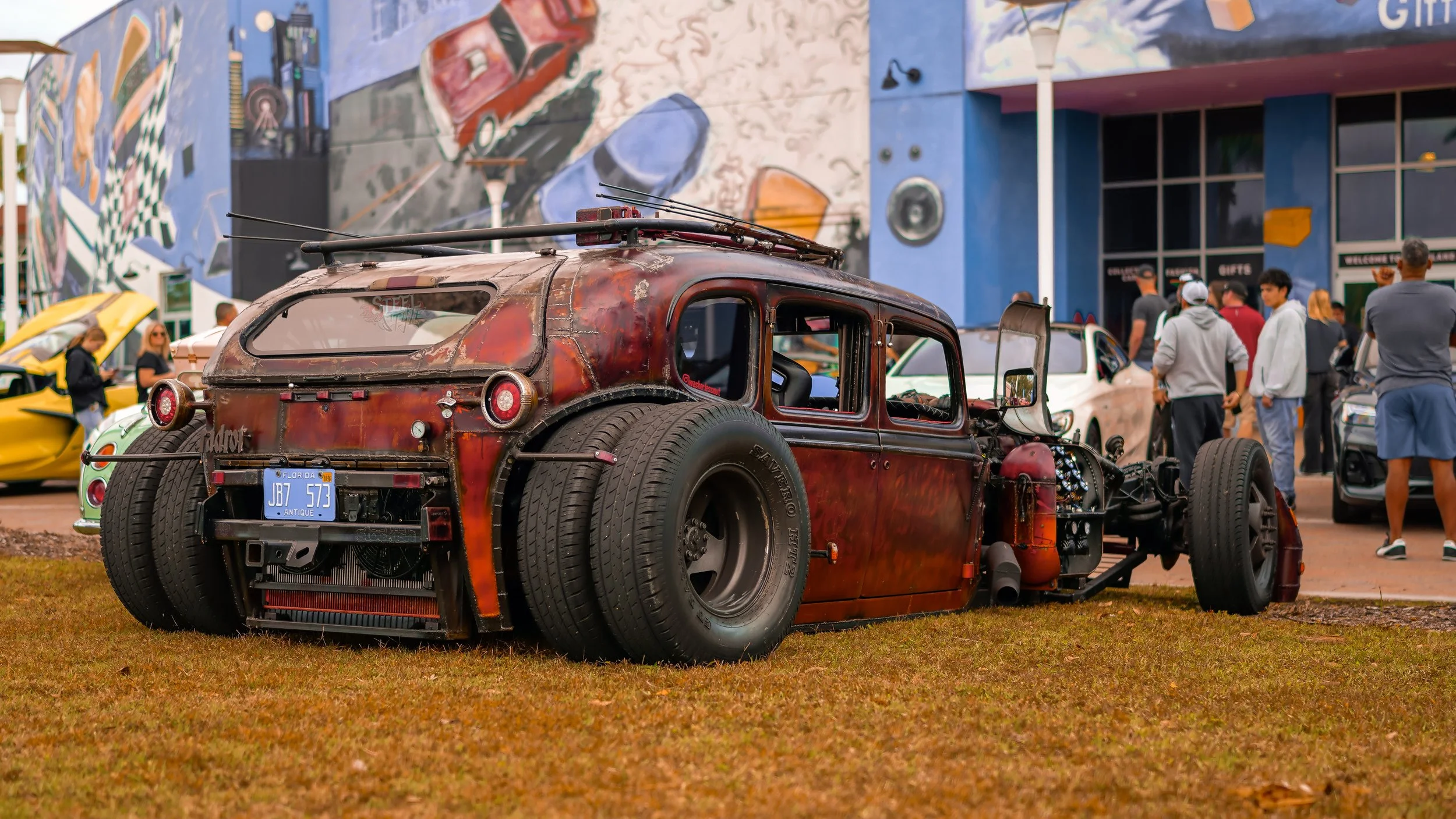 A rusty, partially disassembled vintage car with large tires is on display at a car show, surrounded by people and colorful buildings.