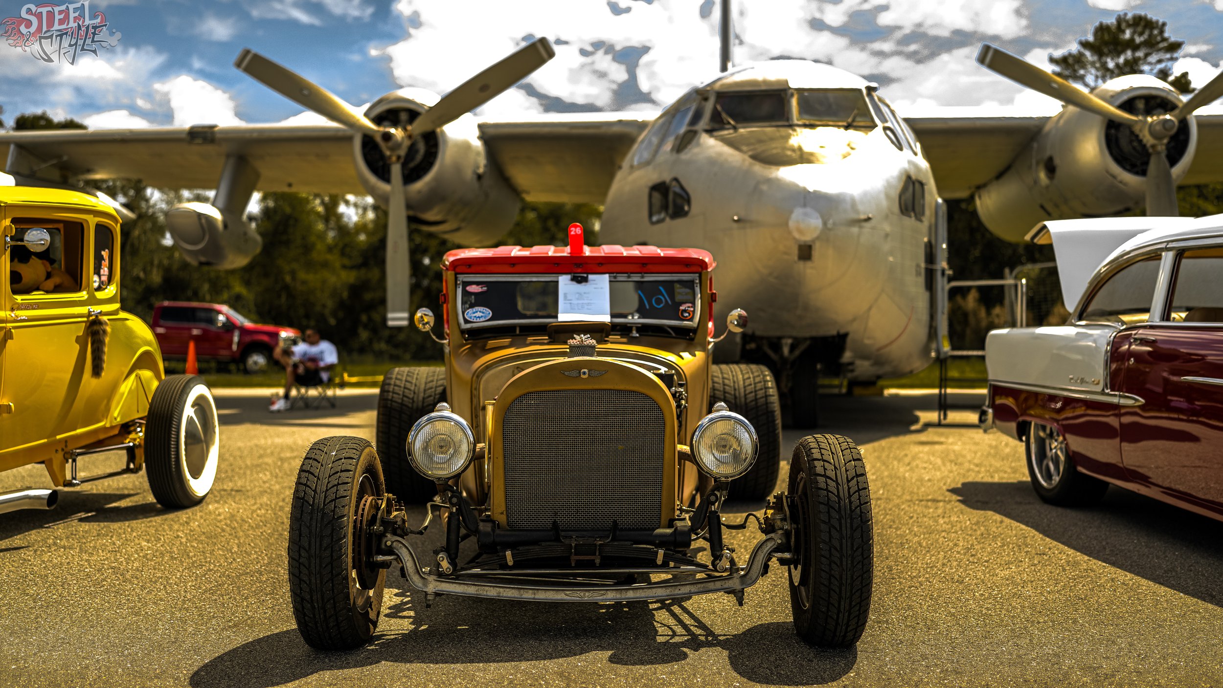 Vintage cars and an airplane on display at an outdoor event, with a blue sky and clouds overhead.