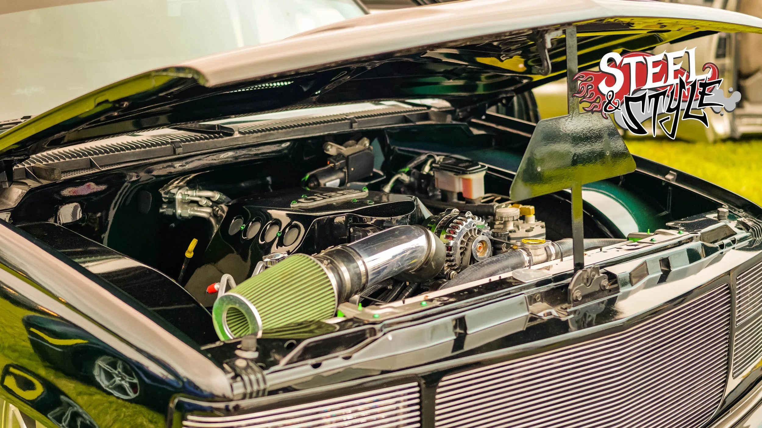 Close-up of a high-performance car engine with a green air filter and shiny metallic components, installed in an open hood, with a sticker that says 'Steel & Style' on the top right corner.