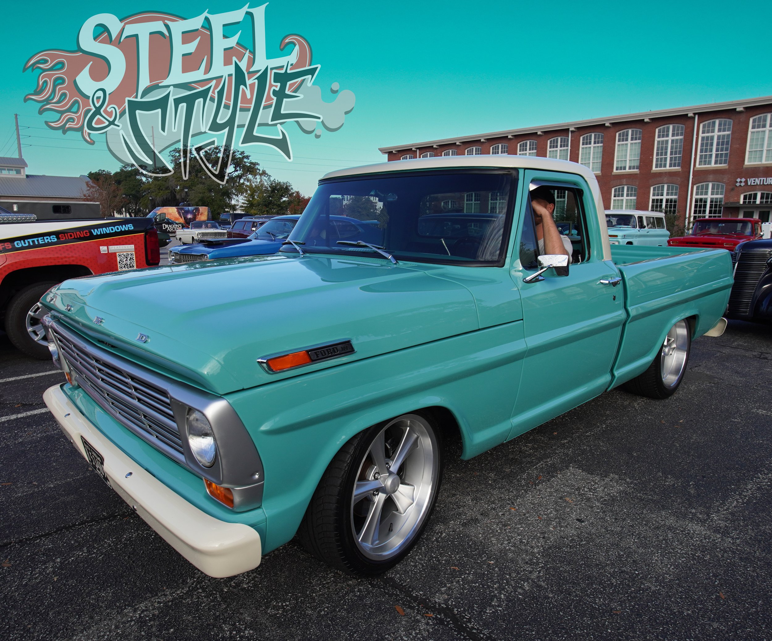 A vintage teal Ford pickup truck parked at an outdoor car show with other classic cars and a brick building in the background.