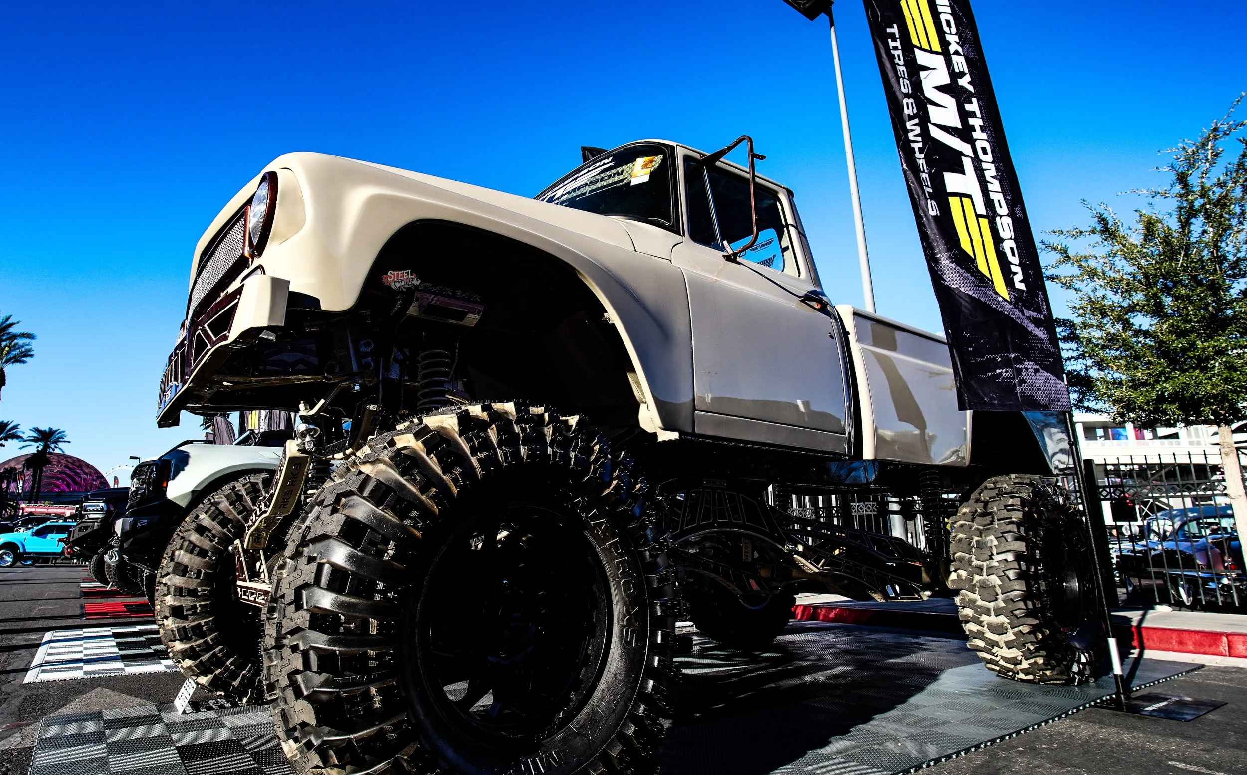 A customized off-road pickup truck with large tires on display outdoors at a motor show under a clear blue sky, with flags and other cars visible in the background.