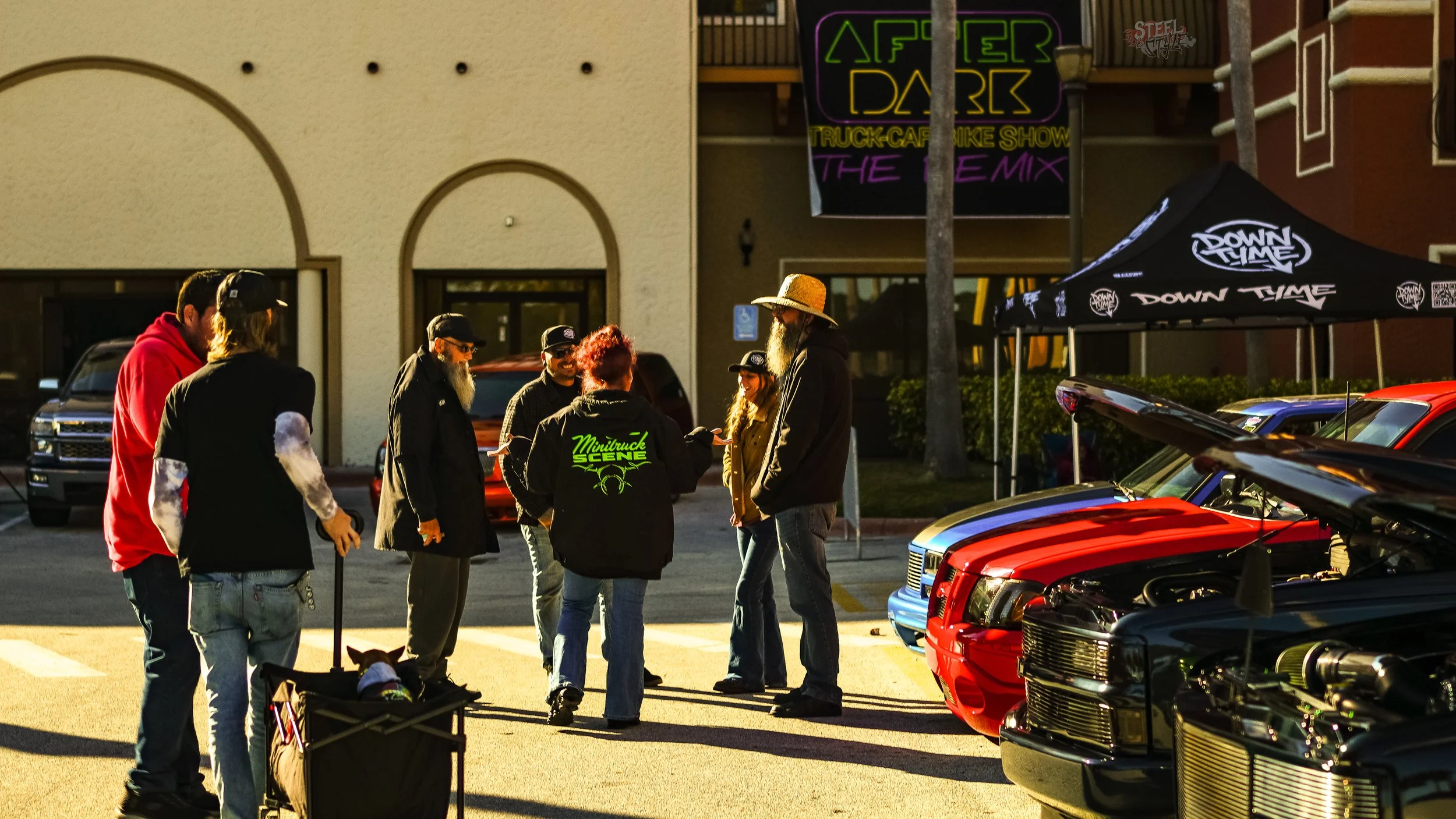 A gathering of people at a car show, with cars on display and a tent with 'Down Time' branding. The group is conversing outdoors, some wearing hats and jackets, with a person pushing a stroller.