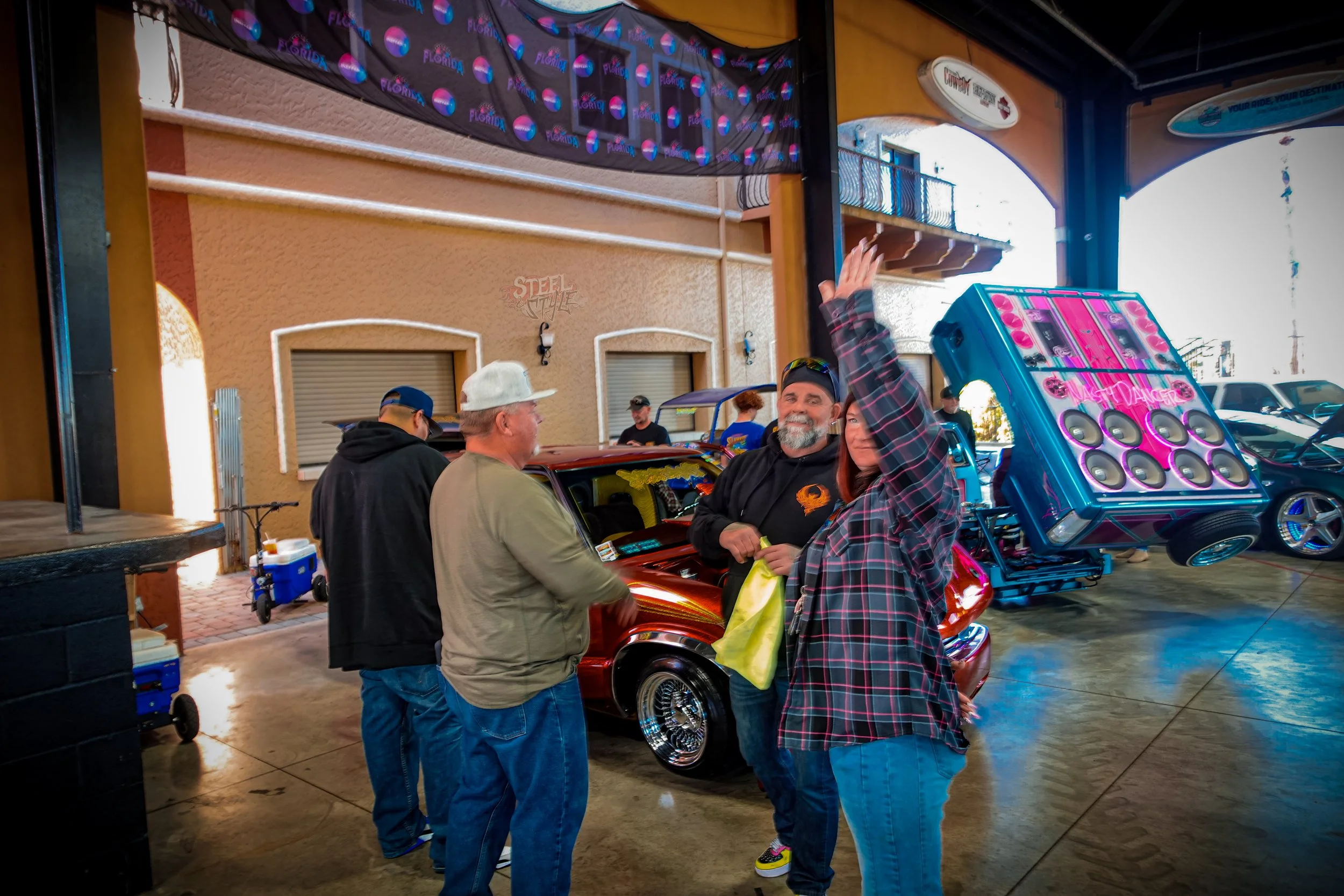 People at a car show, with a woman in a plaid shirt raising her hand, a man with a beard in black clothing, and two men in conversation near a shiny vintage car, in an indoor setting.