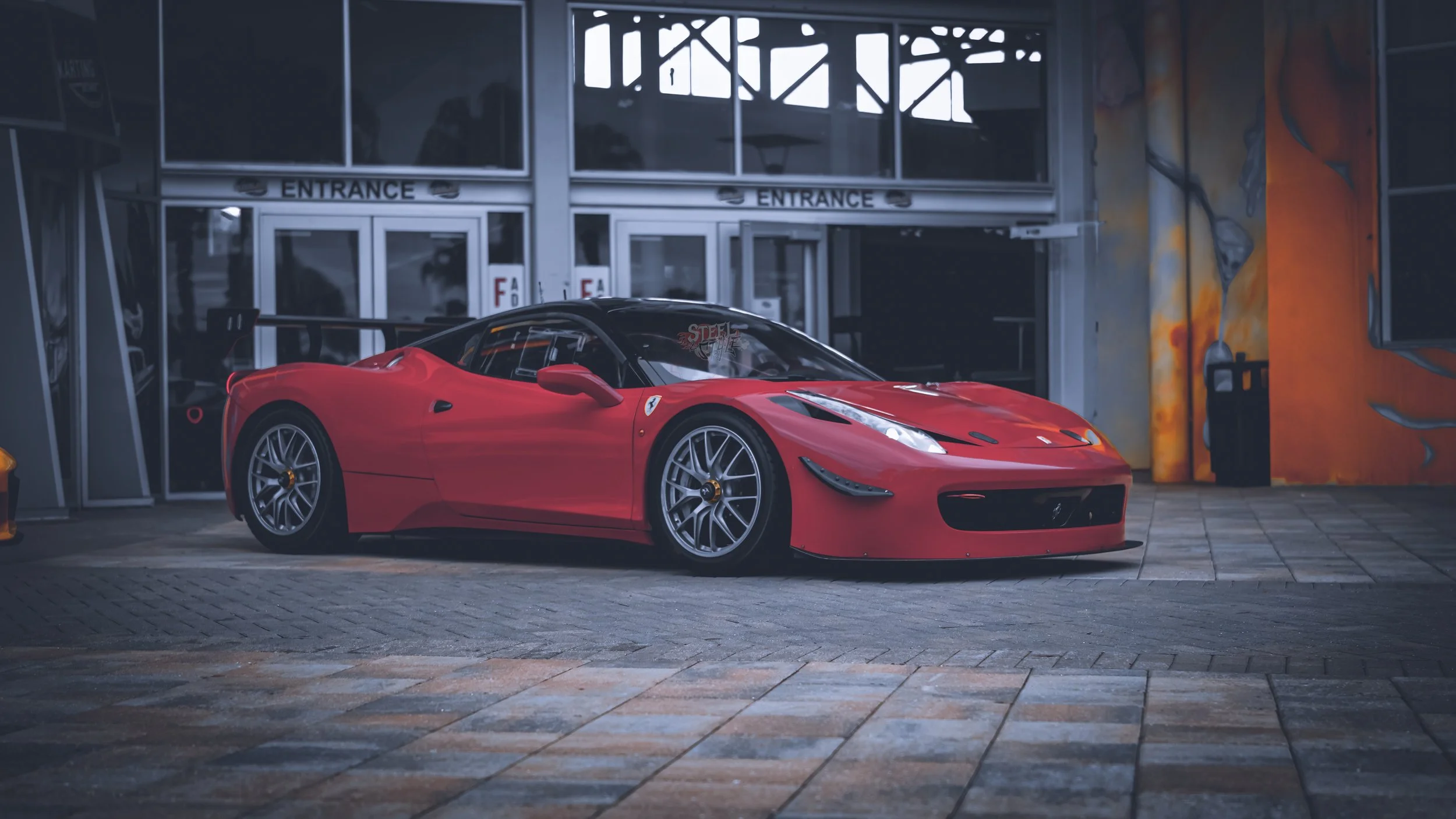Red Ferrari sports car parked outside of a building with glass doors labeled 'Entrance'.