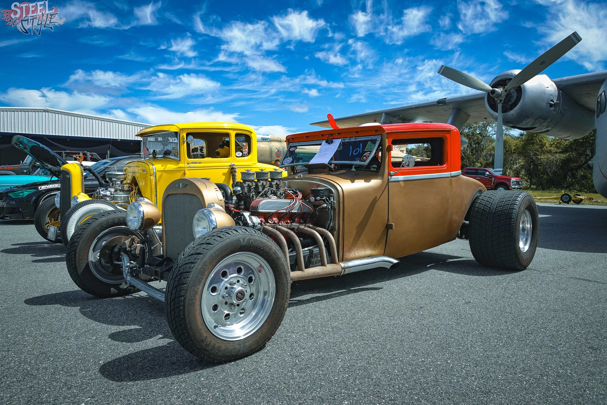A vintage hot rod car with an exposed engine and large tires parked outdoors at a car show, with old aircraft and other vehicles in the background under a partly cloudy blue sky.
