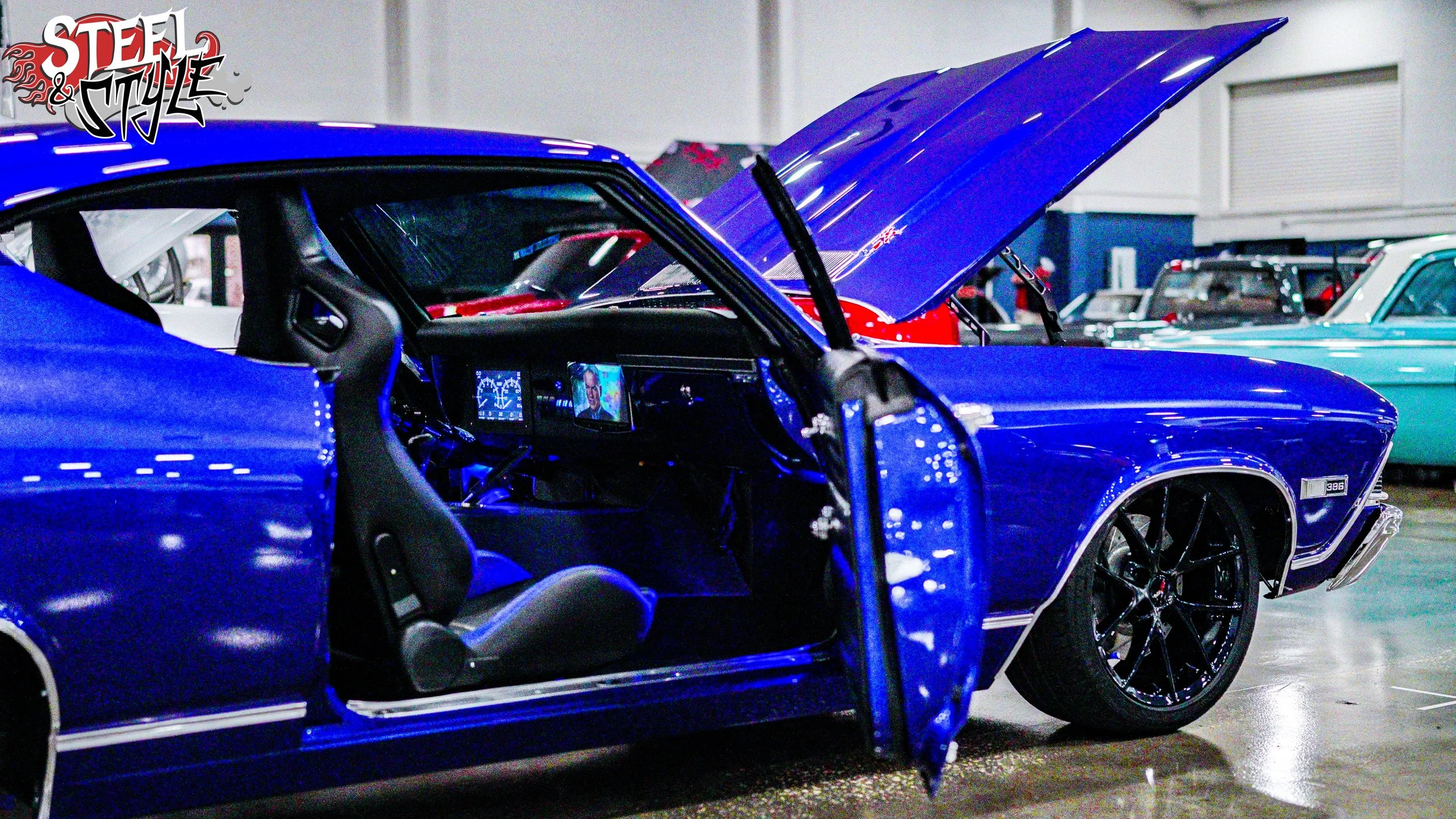 Blue muscle car with its door open at an indoor car show, with an open hood revealing part of the engine, and other classic cars visible in the background.