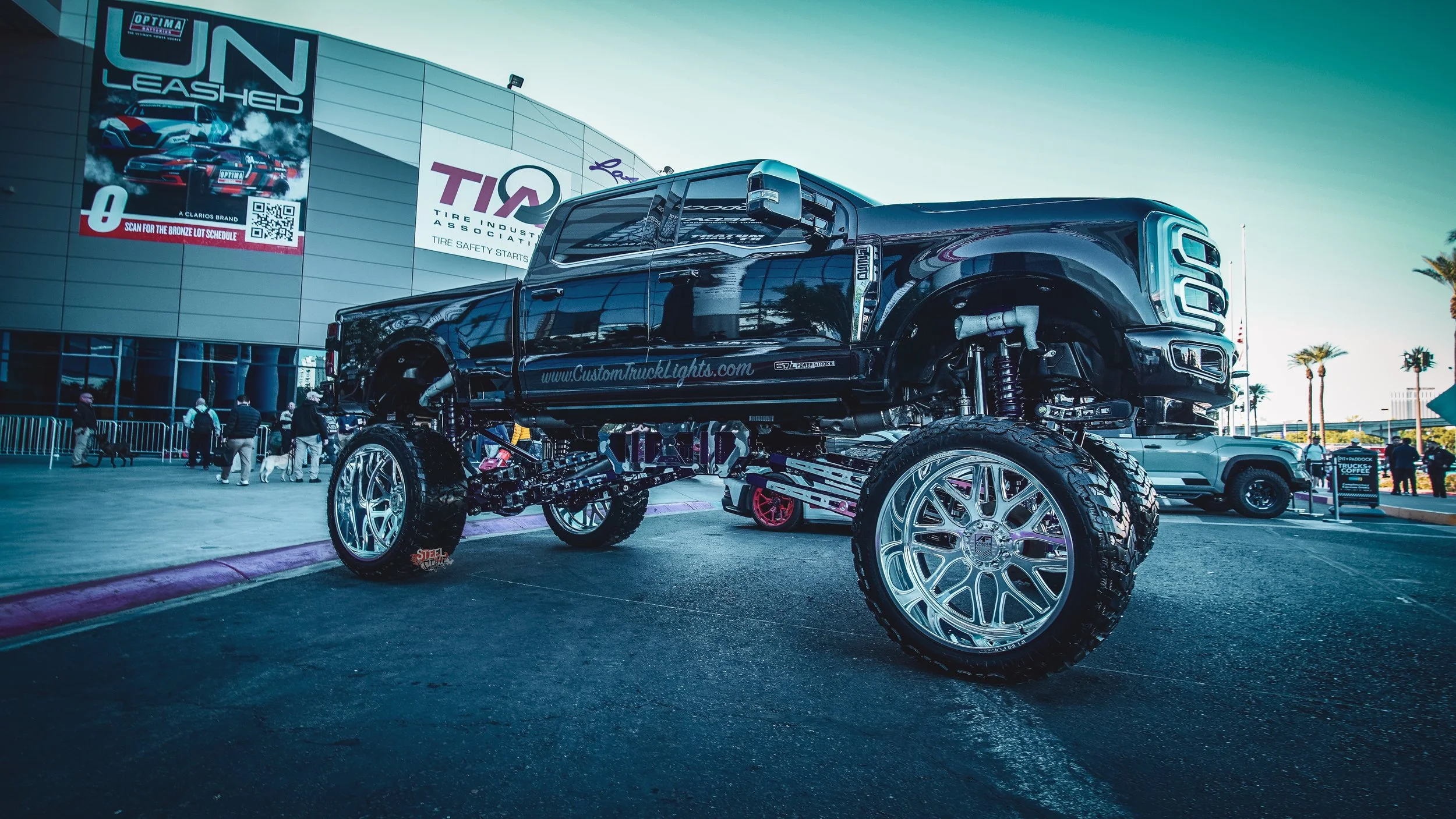A lifted black pickup truck with large chrome wheels on display at an outdoor event in front of a building with advertisements. The truck's undercarriage and suspension are visible, and there are people and other vehicles in the background.