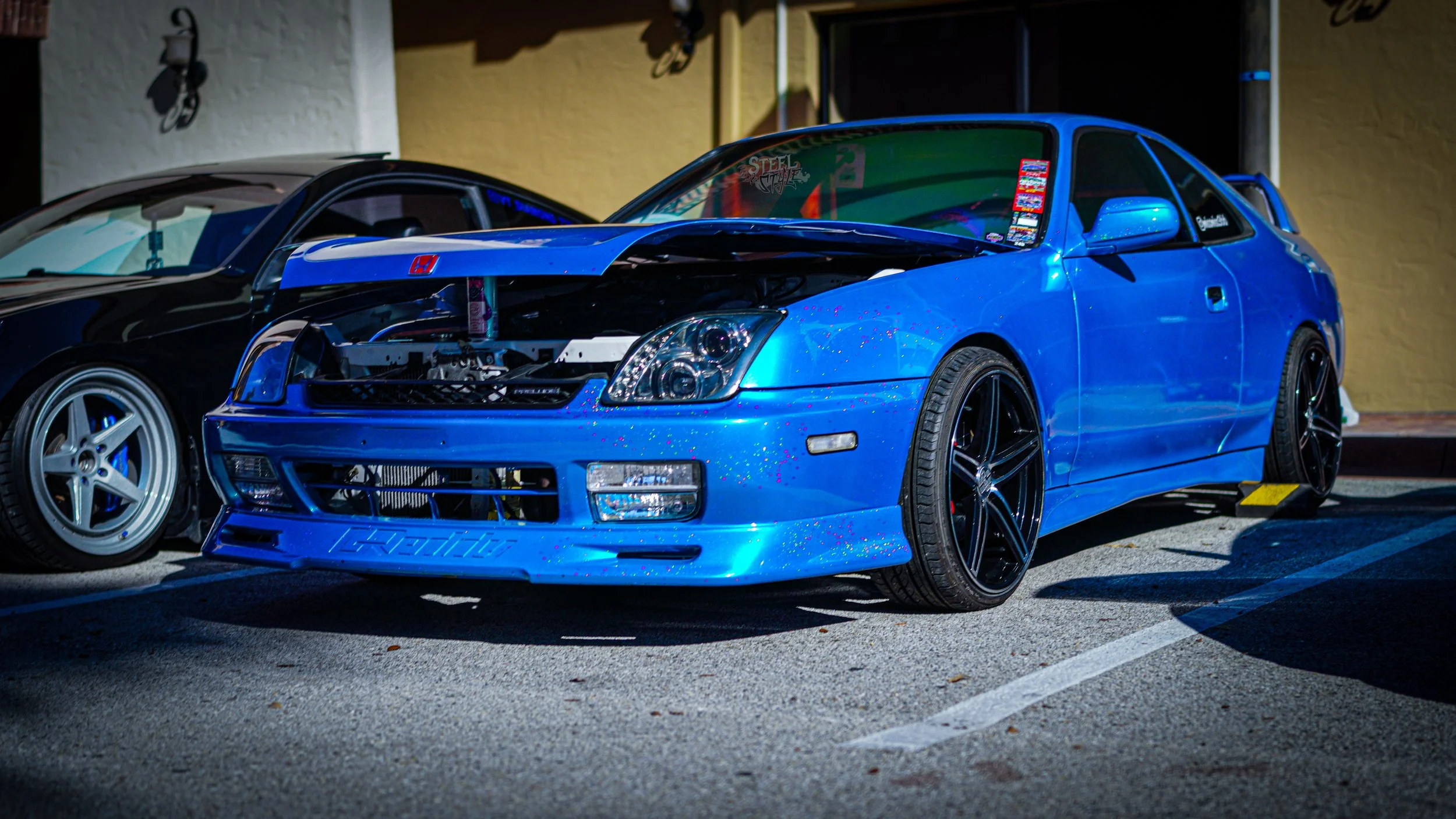 Blue modified sports car with open hood, black aftermarket wheels, parked in a parking lot at night, next to a black car, with a yellow wall and building in the background.