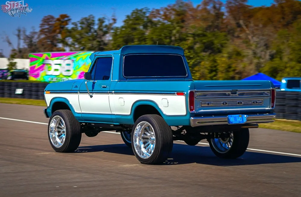 A blue and white vintage Ford pickup truck with large chrome wheels driving on a race track during daytime.